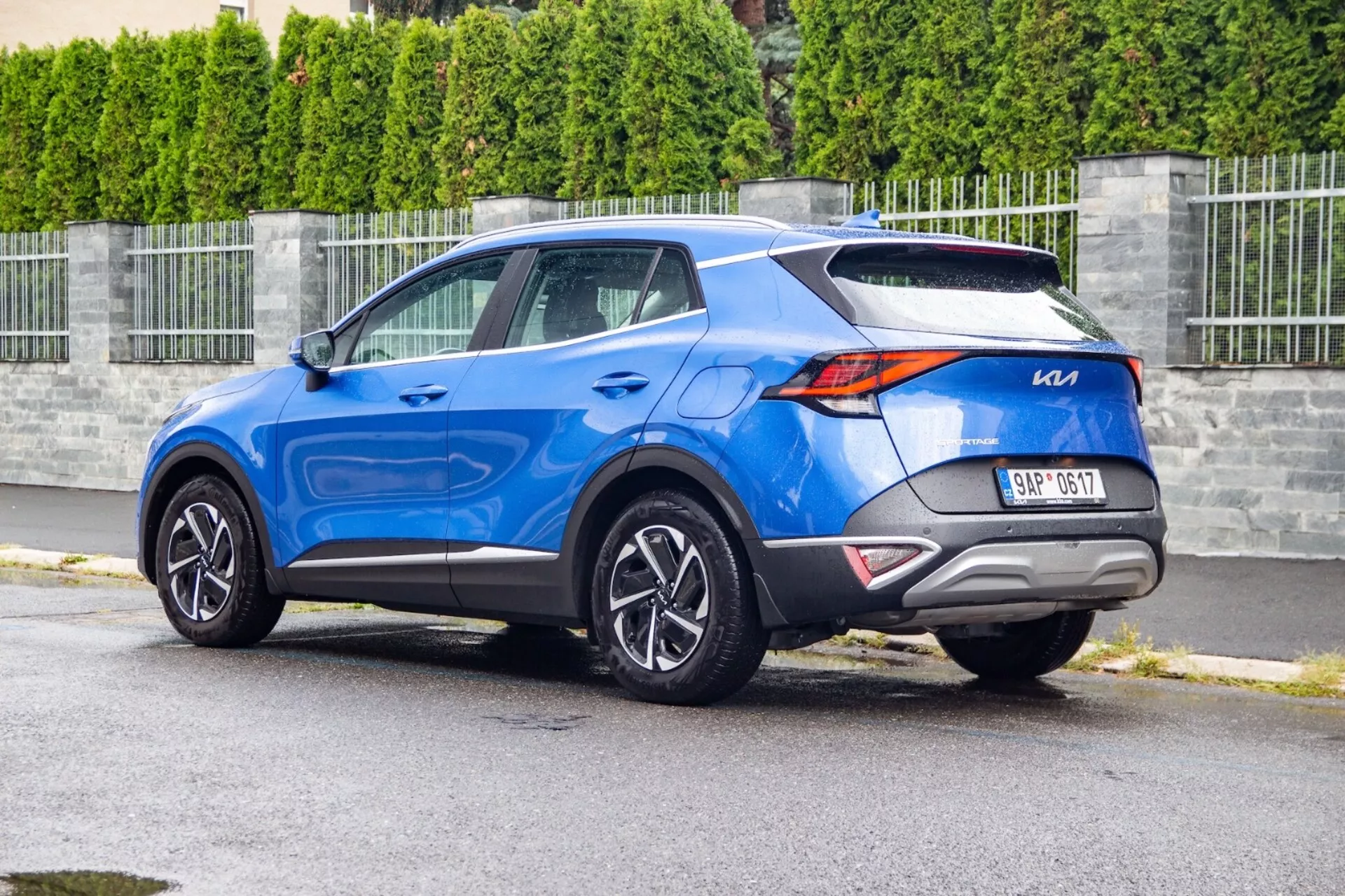 Kompakt SUV Kia Sportage 2022 i blått, som visar LED-bakljus, integrerade parkeringssensorer, dynamisk sidoprofil med stigande midjelinje, flerspakiga legeringshjul, sidospeglar med blinkers, hajfenantenn och takräcken, fångad från ett bakre tre-kvarts vinkel som framhäver design och funktioner