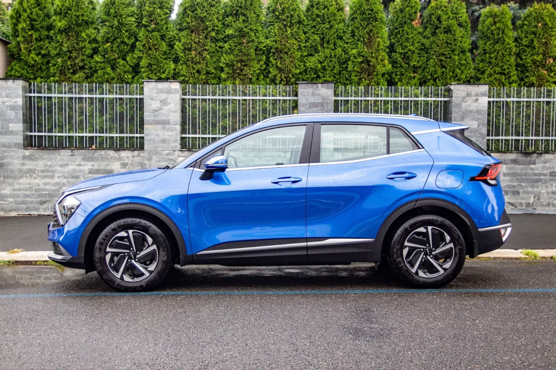 Crossover SUV Kia Sportage 2022 i lysande blått, som visar sin slanka, kompakta design med ett kupé-liknande sluttande taklinje, konturerad huva, LED-strålkastarenheter, fem-ekrade lättmetallfälgar och aerodynamiska förbättringar som en bakre spoiler, i en förortsinställning.