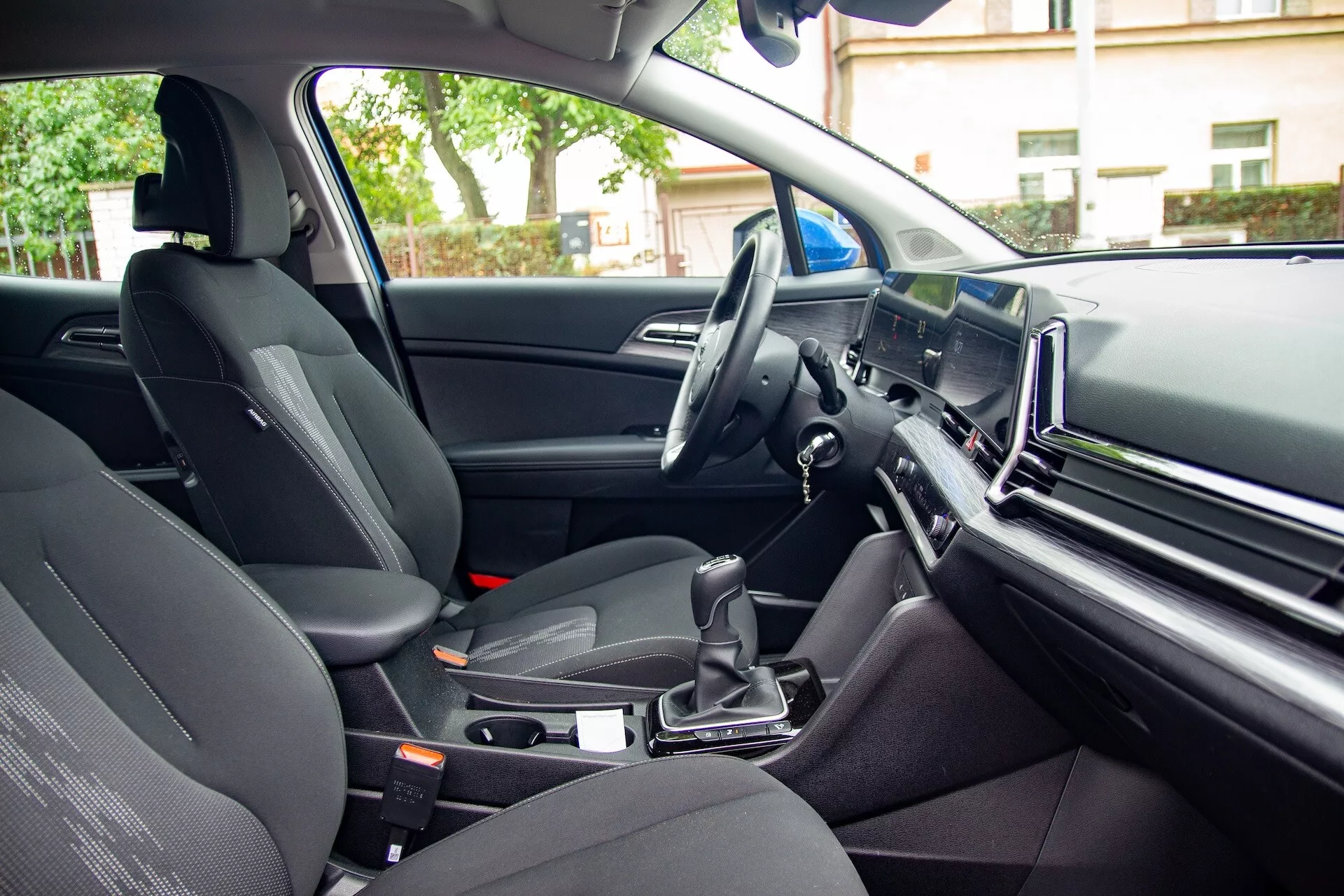 Interiörvy av Kia Sportage 2022 från baksätets passagerares perspektiv som visar framsätets passagerarsäte, mittkonsolen med automatväxellåda, ratten på vänster sida och en stor infotainmentskärm på instrumentbrädan