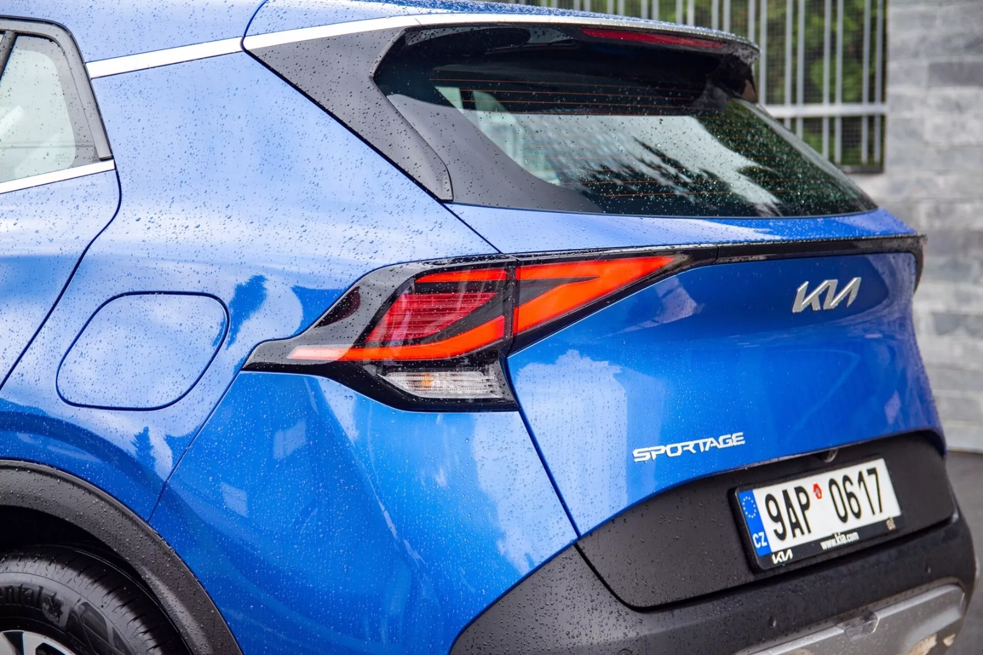 Delvis vy av en blå Kia Sportage 2022 som framhäver de sofistikerade Z-formade LED-bakljusen och detaljerna i bakdesignen, inklusive modellmärket, registreringsskylten och regndropparna på lacken