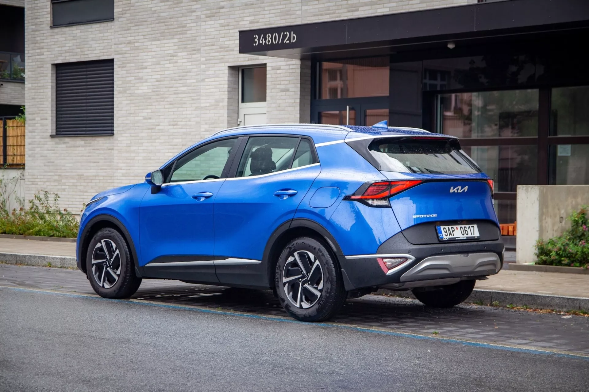 Modern crossover SUV Kia Sportage 2022 parkerad på en stadsgata, visar bakifrån trekvarts vy med distinkta bakljus, sportigt takspoiler, flerspakiga legeringshjul, livligt blå färg och potentiella moderna tekniska funktioner.
