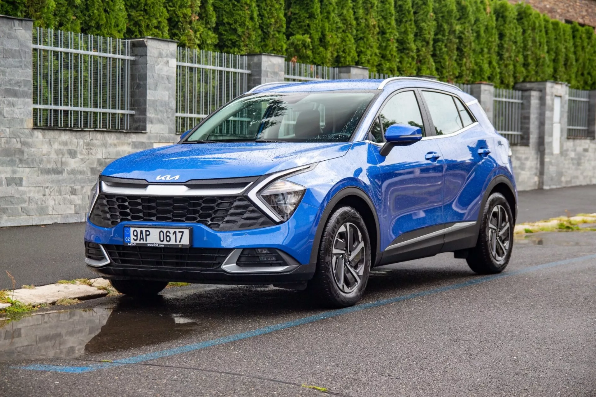 2022 Kia Sportage kompakt SUV i blått parkerad på en våt asfaltgata som visar frontgrill, strålkastare, stötfångare, motorhuv och förarsidans profil från ett trekvartsfrontvinkel med lågt perspektiv för att framhäva fordonets design och teknologiska egenskaper