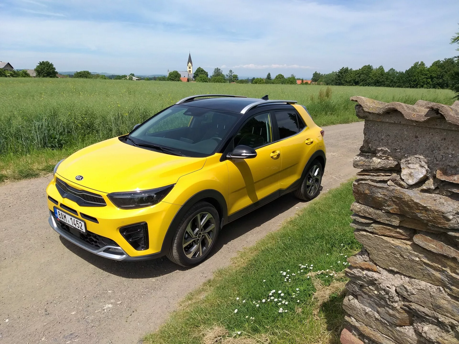 Gul Kia Stonic parkerad på en oasfalterad landsväg, visar sina fram- och sidoprofiler med synlig frontgrill, strålkastardesign och takräcken mot en bakgrund av gräs, små vita blommor och en torr stenmur