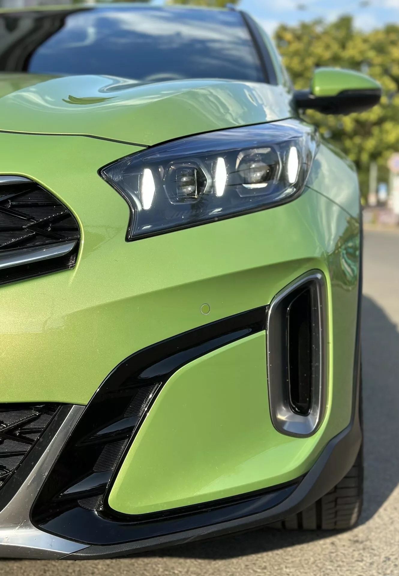 Närbild av 2022 Kia Xceed som visar främre vänster kvartal med detaljerad vy av LED-strålkastarasamlingen, del av grillen, luftintag på frontstötfångaren, inställd mot en limegrön karossfärg, betonar fordonets sportiga design och avancerad belysningsteknologi.