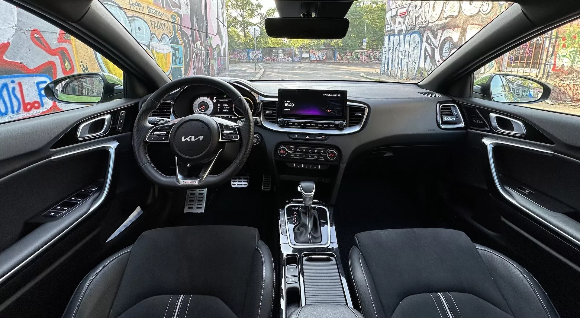 Interiörvy av Kia XCeed 2022 från baksätena, som visar styrratt med kontrollknappar, digital instrumentpanel, centralt infotainment-skärm, manuella klimatkontroller, framstolar klädda i läder, växelspak i parkeringsläge och avancerade funktioner som körlägeskontroller och automatiskt dimmigt backspegel, allt mot en mörk färgsättning.