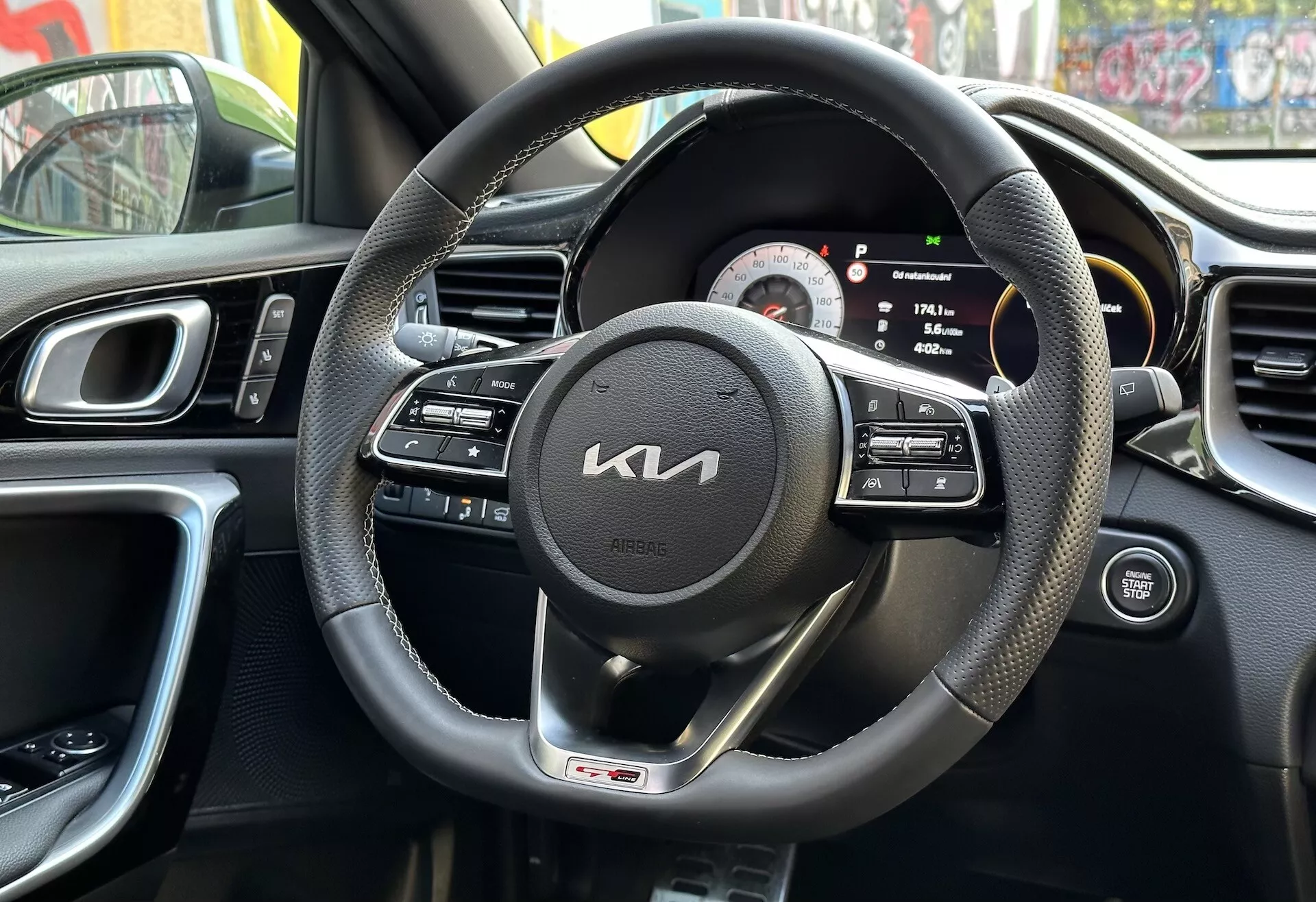 Interiörvy av Kia XCeed 2022 från förarens perspektiv med svart läderklädd ratt med multifunktionskontroller och GT-linjeemblem, digital-analog instrumentkluster, del av mittkonsolen med luftventiler, ergonomisk design av instrumentbrädan, och förarens dörrpanel med kontroller, reflekterande urban miljö
