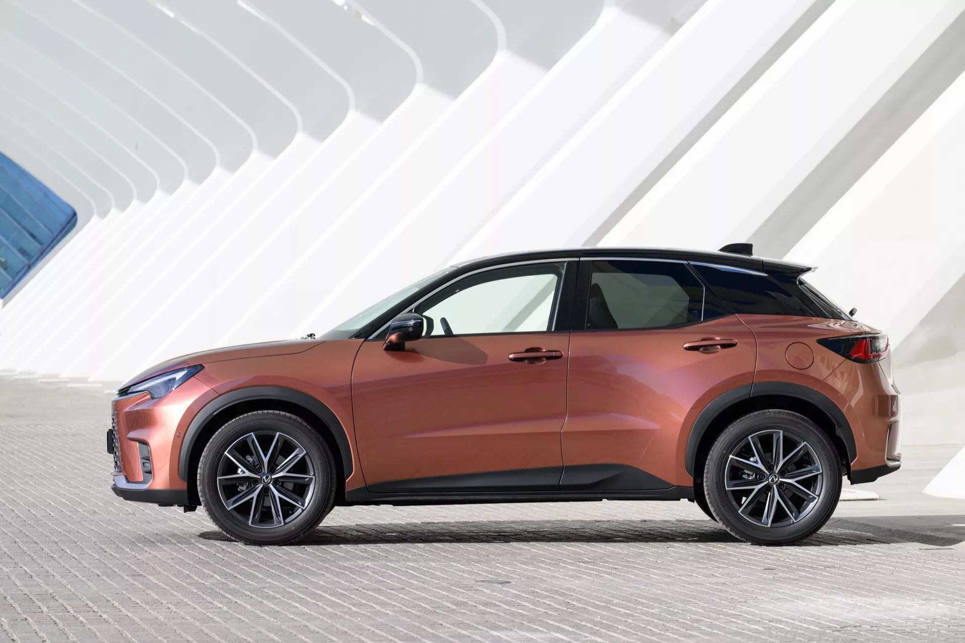 Sidoprofil av Lexus LBX 2024 under konstgjord belysning, framhäver metallisk kopparfärg, eleganta LED-strålkastare och geometrisk bakgrundsstruktur