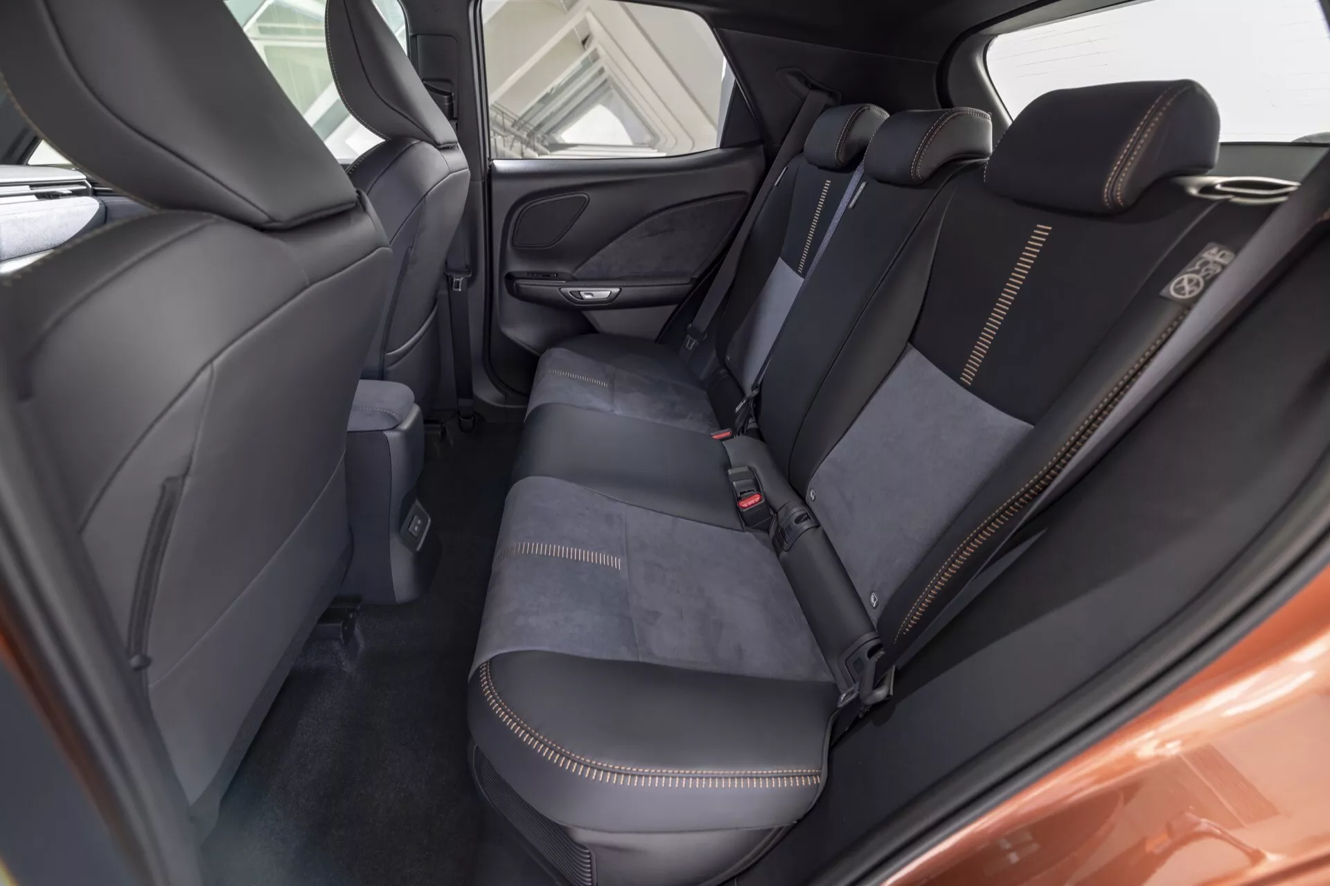 Interiörvy av Lexus LBX 1:a generationen 2024 från bakre passagerarutrymmet som visar baksätesområdet, detaljer i klädseln med vit kontrastsöm, quiltade ryggstöd, fällbart armstöd, dörrhögtalargaller och förvaringsficka på framsätets baksida