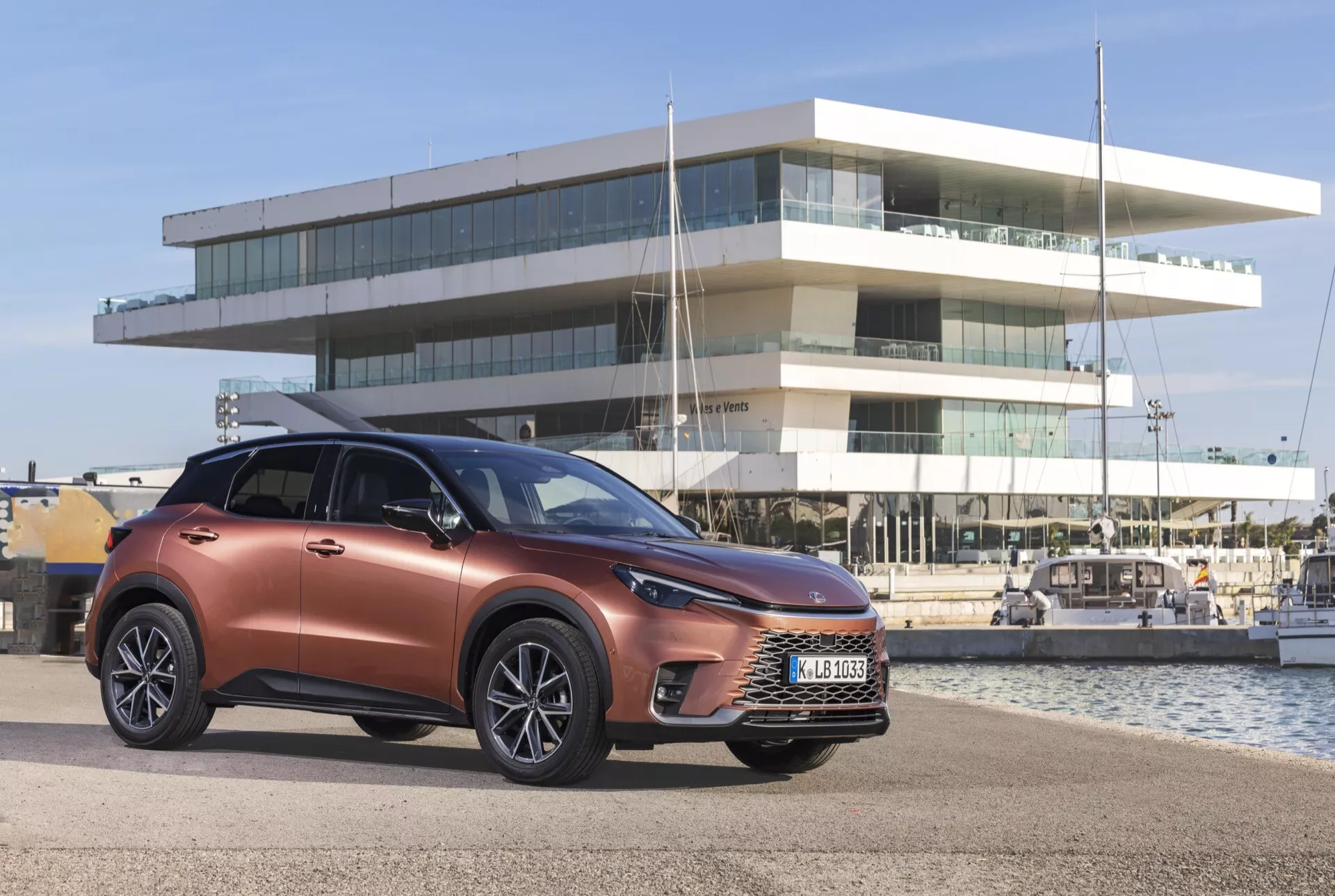 2024 Lexus LBX 1:a generationens moderna crossover SUV i metallisk koppar, visar fram grill, LED-strålkastare och legeringshjul, placerad utomhus med en marina och en samtida byggnad i bakgrunden, vilket betonar dess inriktning mot en urban/kustnära livsstil.