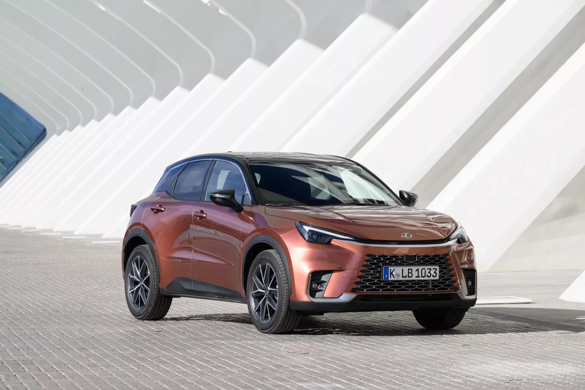 Lexus LBX av första generationen 2024 visas i dagsljus, med en distinkt frontgrill och slanka LED-strålkastare, mot en bakgrund av modern arkitektur med återkommande vinklade geometriska former. Vinkeln framhäver bilens aerodynamiska kaross, metallfärg och legeringshjul, betonande dess crossover SUV-design och avancerad fordonsingenjörskonst.