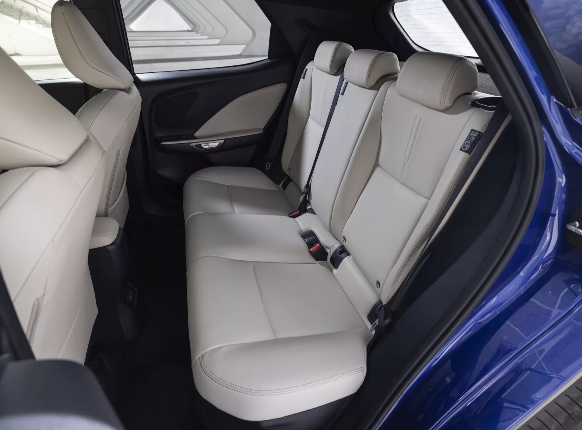 Interiören i Lexus LBX 1:a Generation 2024 visar lyxiga ljusbeige lädersäten bak med huvudstöd, säkerhetsbälten, dörrpanel med kontroller, bakre delen av mittkonsolen inklusive luftventiler och laddningsportar, under välupplysta förhållanden som betonar komfort och säkerhetsfunktioner