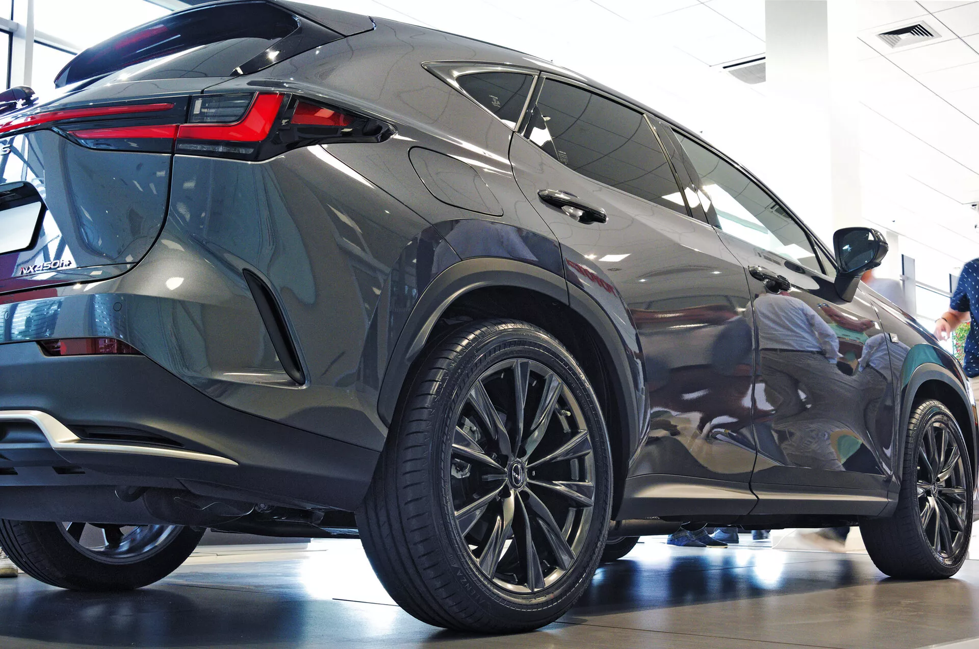 Treboksvinkel bakifrån på Lexus NX 2021 SUV som visar passagerarsidans profil och den bakre delen med stora mångekrade lättmetallfälgar, lågprofildäck, skarpa bakre LED-ljus, metallisk kaross, tonade rutor, integrerad bakre spoiler och polerad showroommiljö som betonar modern estetik och lyxiga funktioner