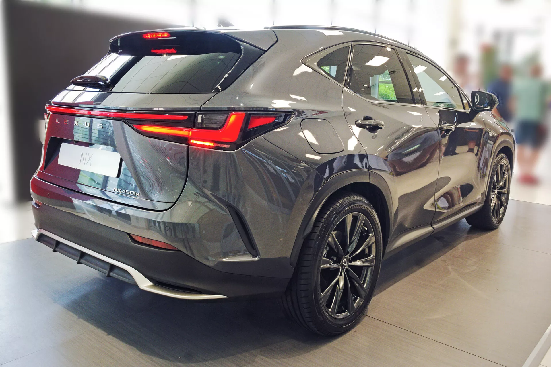 Bild som fångar Lexus NX 2021 från en bakre trekvarts vinkel, som visar dess eleganta och aerodynamiska konturer, LED-bakljus, skarpt lutande bakruta, bakspoiler, bakrutetorkare, tvåfärgad bakre stötfångare med parkeringssensorer, diffusor-stilelement, flerspoksfälgar och dess metalliska grå färg mot det polerade golvet i inomhusutställningshallen.