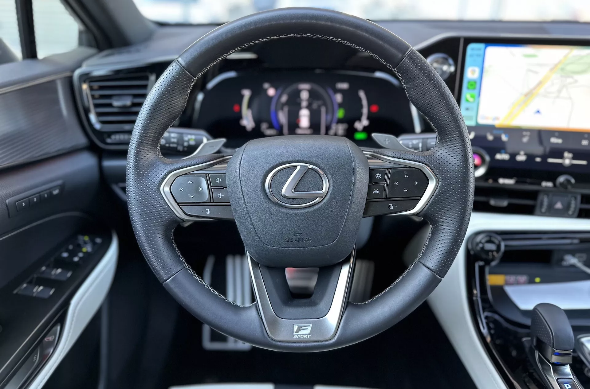 Interiören i Lexus NX 2021 med fokus på det läderomslutna, multifunktionella ratten med Lexus-emblem och F SPORT-logotyp, digital instrumentpanel som indikerar hybrid drivlina, samt en glimt av GPS-navigation på underhållningssystemet