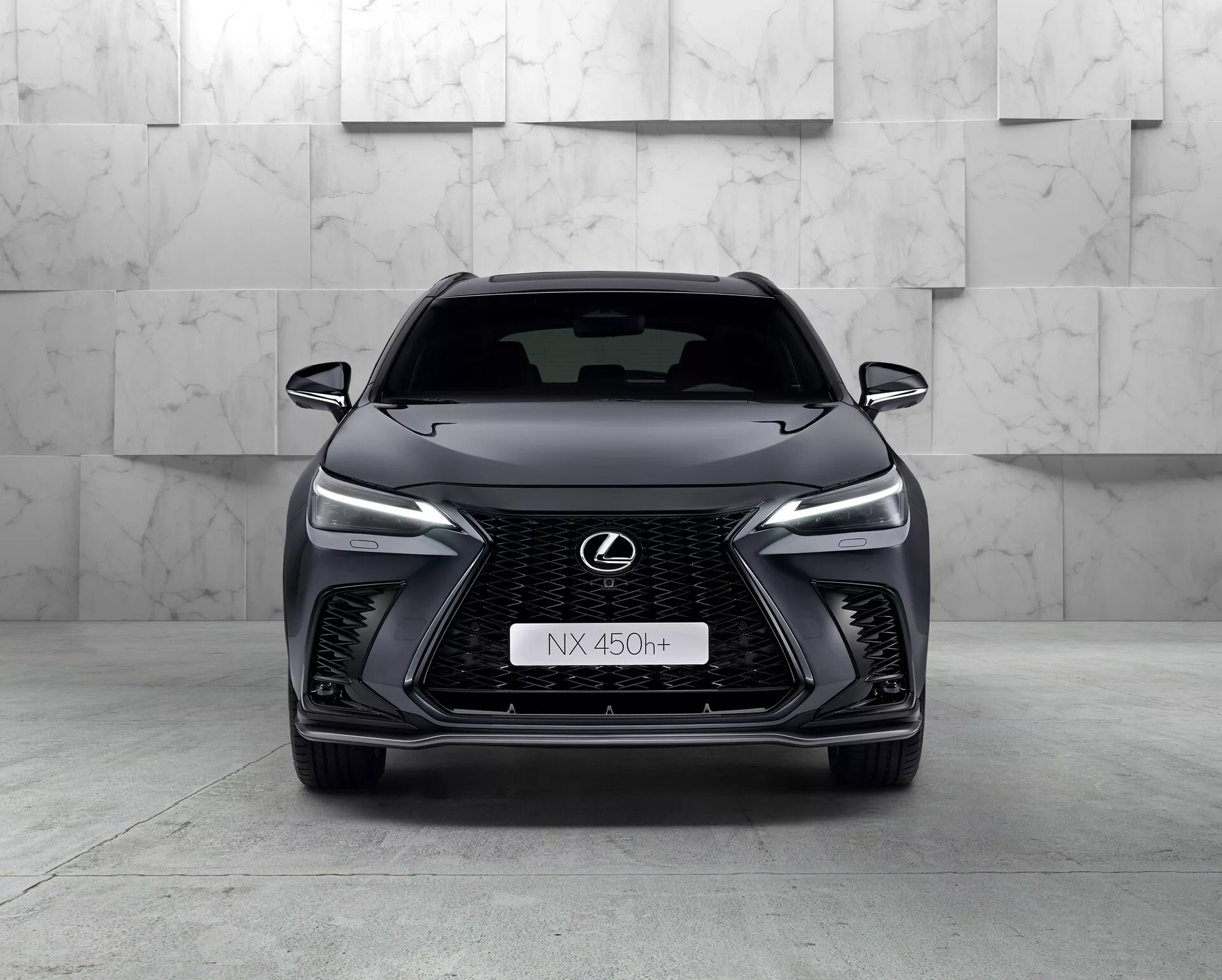 Frontal vy av 2021 Lexus NX 450h+, en elegant modern SUV som visar sin framträdande spindelformade grill med nätmönster, smala LED-strålkastare, kromad Lexus-emblem med blå kontur och aggressiv främre stötfångardesign. Bilens hybridkaraktär anges av 'h+'-emblemet på grillen. Studiomiljön framhäver fordonets kurvor och egenskaper med en ren, kontrollerad belysning.