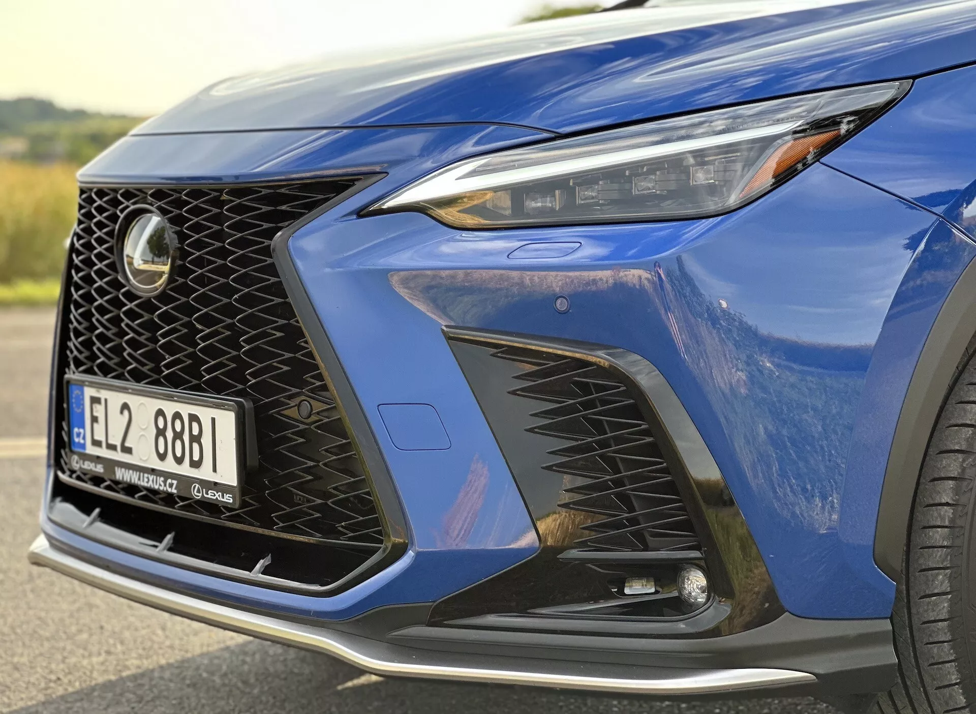 Närbild på främre hörnet av Lexus NX 2021 som visar strålkastaren med LED-teknik, distinkt nätdesigngrill och aerodynamisk frontstötfångare med luftintag, framhävd av en djupblå metallisk färg och en glimt av ett lyxigt legeringshjul.