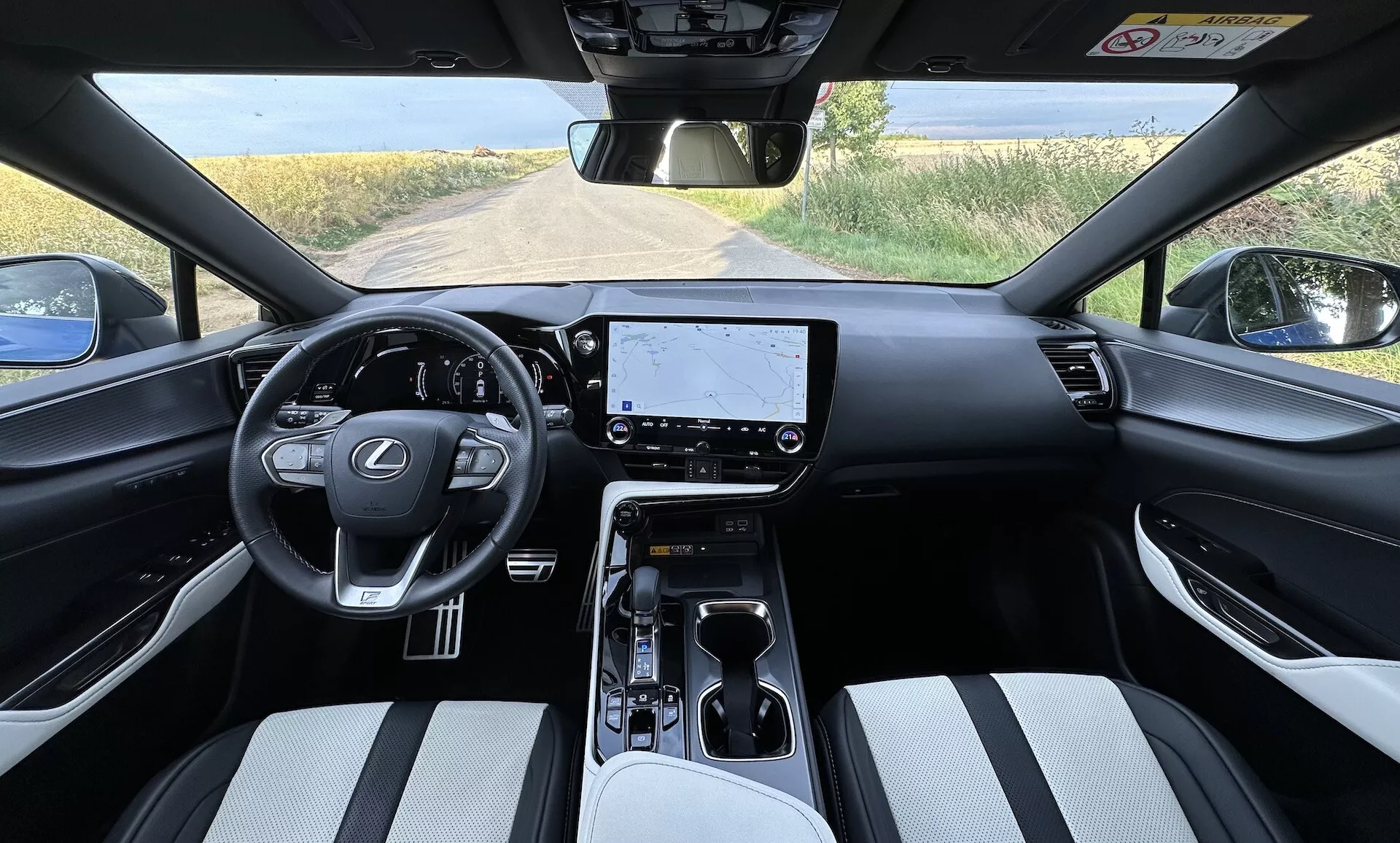 Interiör vy av Lexus NX 2021 från förarens perspektiv med en centralt placerad ratt med Lexus logotyp och kontrollknappar, en stor infotainment pekskärm på instrumentbrädan, minimalistiska kontroller för klimatanläggningen, en växelväljare för automatisk växellåda på mittkonsolen med mugg hållare och förvaring, tvåfärgade klädsel på sätena, och en delvis utsikt över ett landskap genom vindrutan.