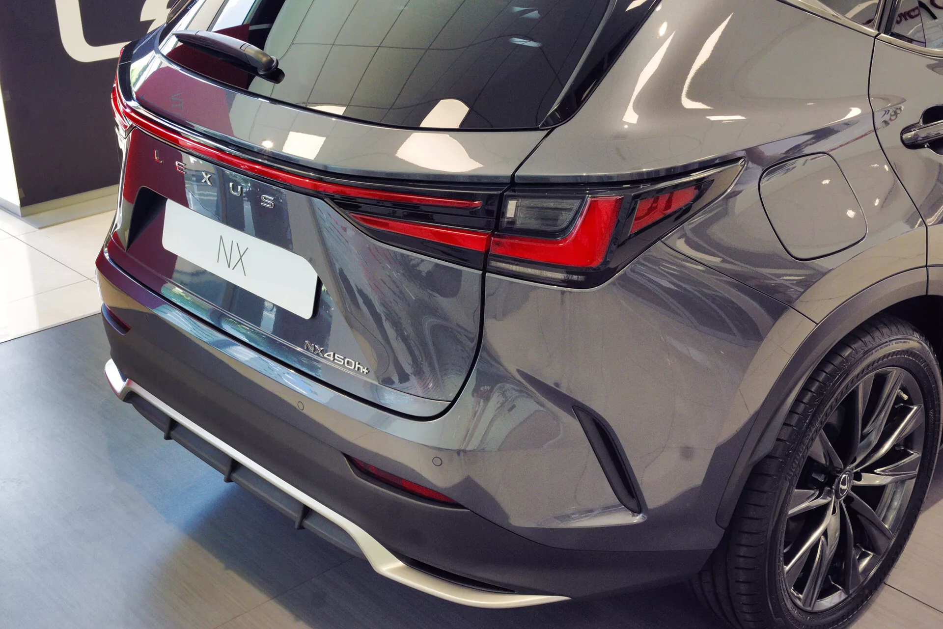 Denna bild visar en modern SUV från en trekvarts bakåtvinkel, vilket framhäver fordonets bakre ände. Bilen är parkerad med högra bakre sidan närmare kameran, vilket ger en klar vy över funktioner som takspoiler, bakruta med torkare och den slående horisontella LED-ljusstången som sträcker sig över fordonets bredd. Tillverkarens emblem och modellbeteckning är tydligt visade på den nedre delen av bakluckan, och det angivna registreringsskyltområdet är omgivet av en kromlist. Den bakre stötfångaren sticker utåt och rymmer kanske parkeringssensorer och en parkeringskamera. Metalliclacken lyser under showroom-belysning, med designen på den mångekrade fälgen på det bakre hjulet i sikte. Fordonet visas sannolikt i ett showroom, vilket framhäver dess lyxiga SUV-design.