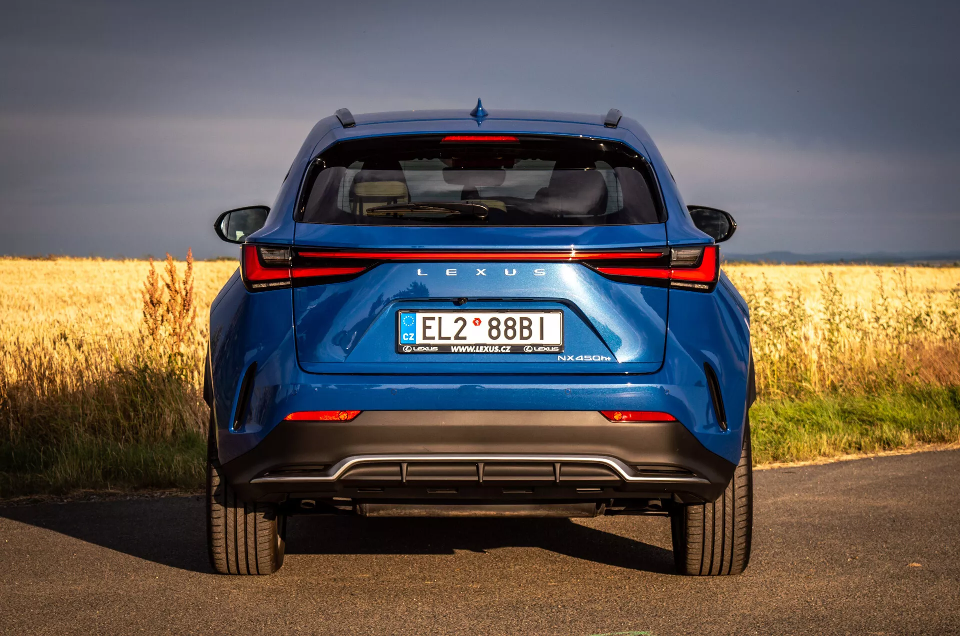 Bakvy av blå SUV Lexus NX 2021 på asfalterad väg med gyllene fält i bakgrunden, framhäver hybridteknologi och designegenskaper