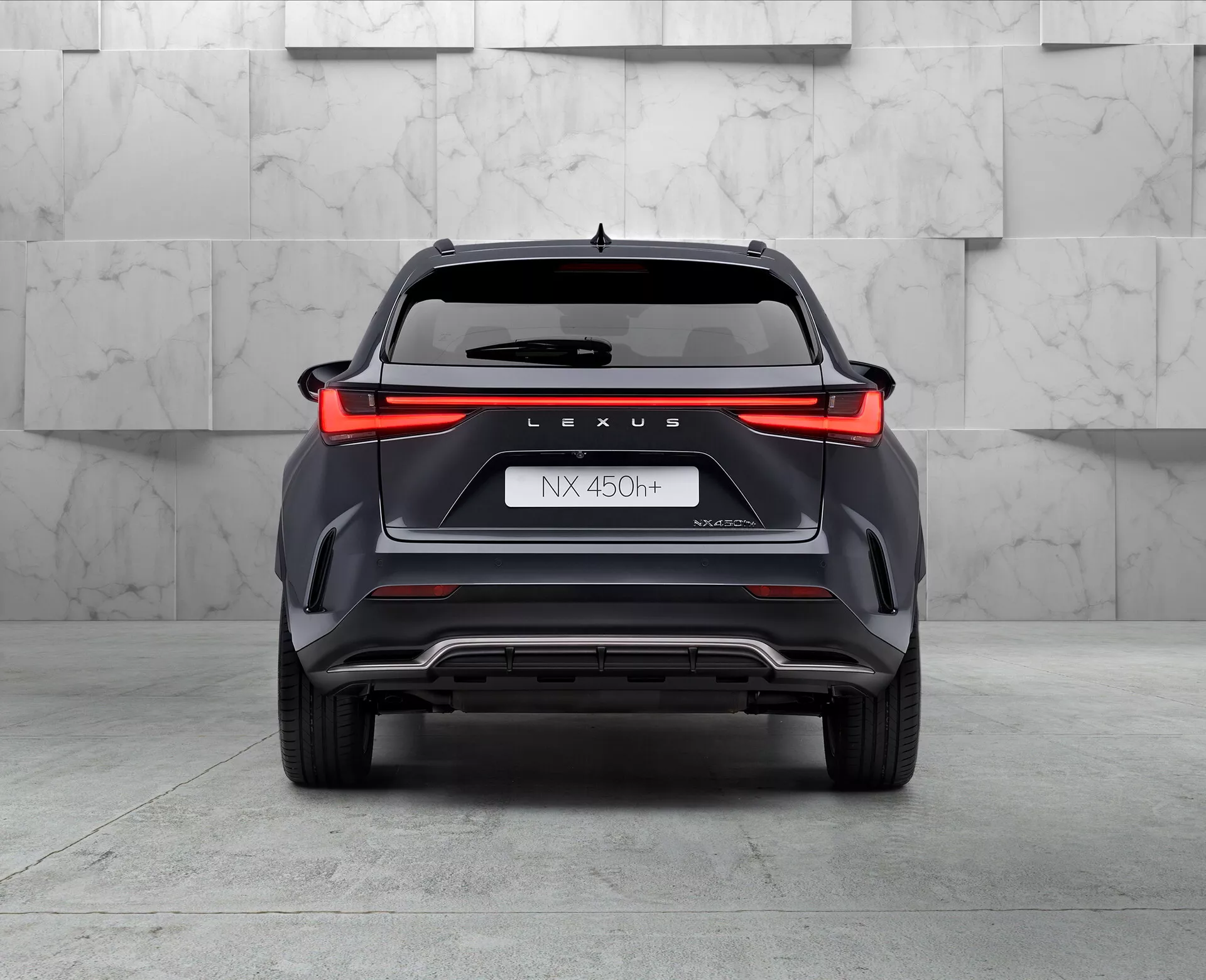 Bakifrånvy av hybrid-SUV:n Lexus NX 450h, fångad från en låg vinkel något skiftad till vänster, som visar bakljusdesignen över hela bredden, 'LEXUS'-märkningen, modellbeteckningen 'NX 450h', namnet 'NEXUS' och hybridsymbolen 'h'. Stötfångare med vinklade stilinslag, ventilationsfunktioner och en silverskidplate, med ett synligt dubbelavgasrör på vänster sida, allt i en showroom-miljö med polerat golv och en neutral bakgrund.