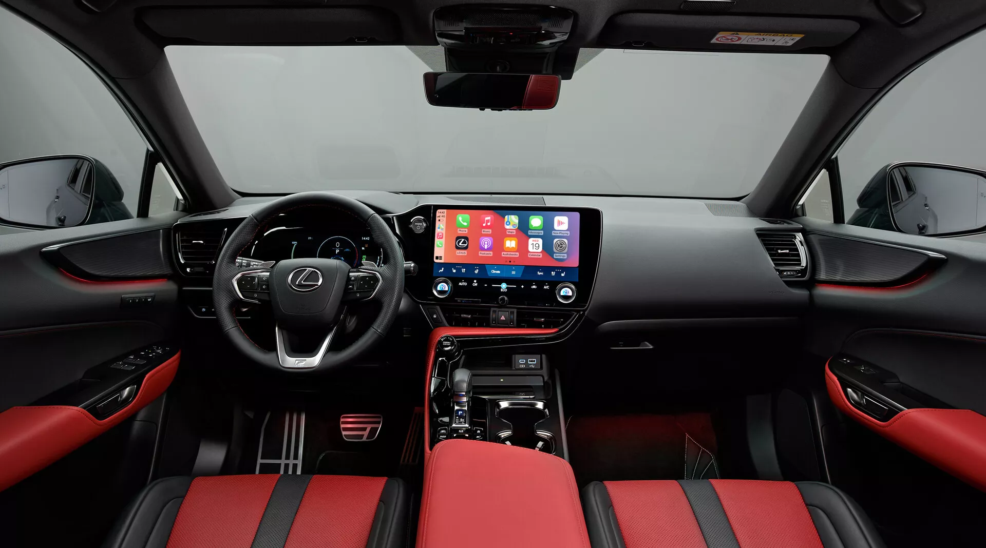 Interiör av Lexus NX 2021 med fokus på den främre kabinen, tagen från en centralt monterad position. Det breda perspektivet visar instrumentbrädan med en stor central infotainmentskärm som visar ett mobilt gränssnitt, fysiska klimatkontroller och en växelspak för automatväxellåda. Den multifunktionella ratten med kontrollknappar antyder avancerade funktioner. Sätena är klädda i tvåfärgat rött och svart läder, vilket ger ett sportigt utseende. Dörrpanelerna följer det röd-svarta temat med kontroller för elhissar och ett synligt högtalargaller, vilket betonar modernitet och lyx.