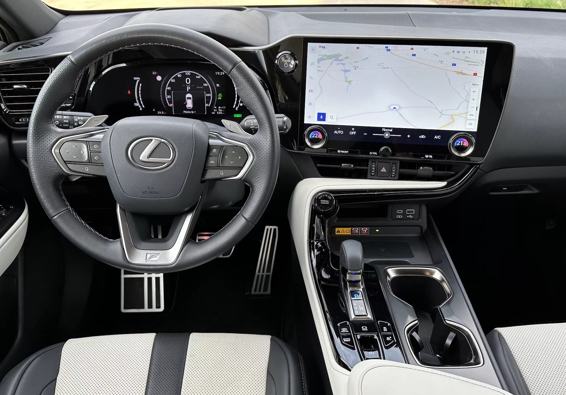 Interiörvy av Lexus NX 2021 som visar förarområdet och mittkonsolen från en höjd vinkel, med Lexus-logotypen på ratten med monterade kontroller, ett upplyst digitalt instrumentkluster, en stor pekskärmsinfotainmentsystem med GPS-navigering, knappar för klimat- och ljudkontroll, en ergonomisk växelväljare, kontroller för körläge, lyxmaterial på säten och kontaktpunkter, metalliska accenter och synliga mugghållare och förvaringsfack.