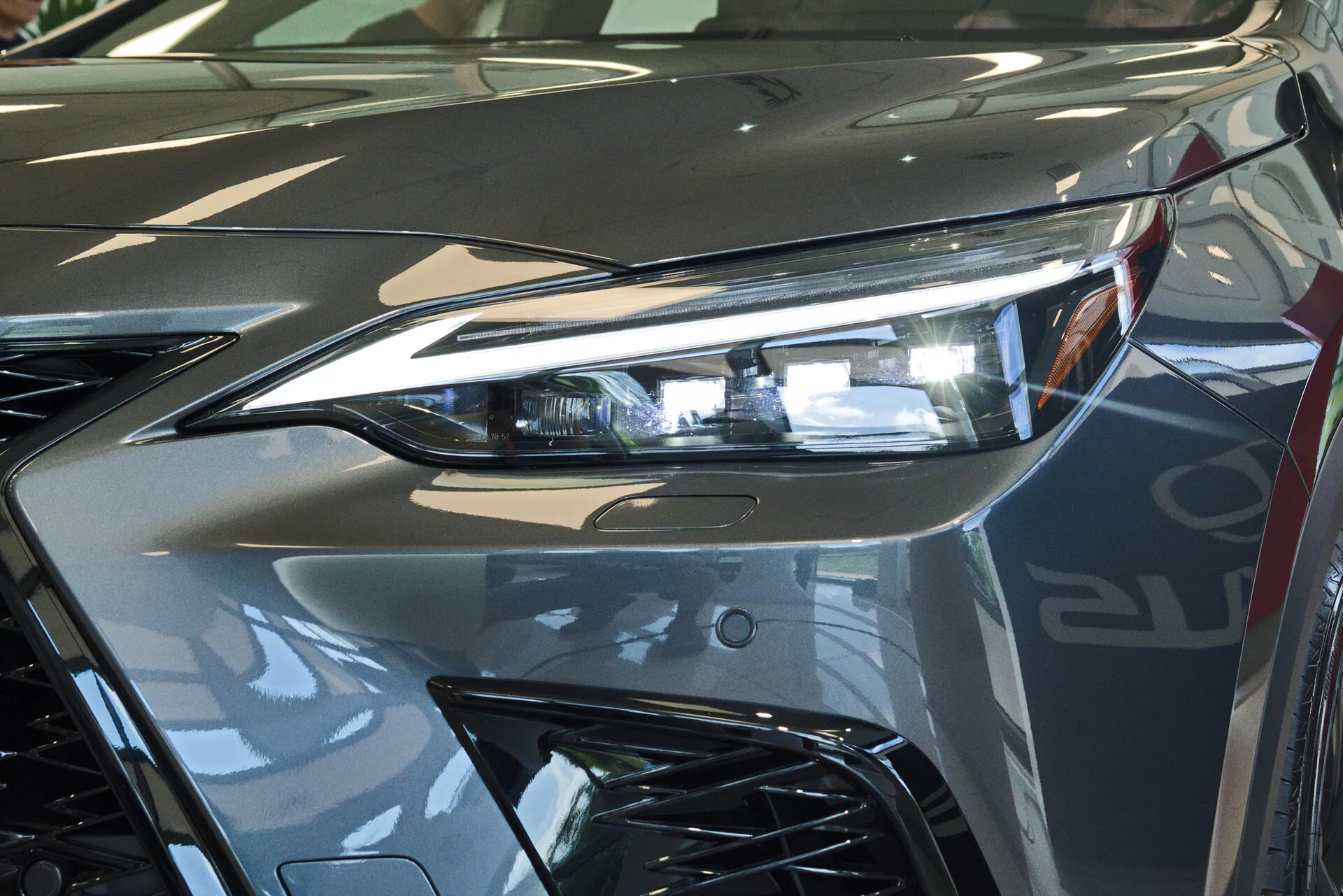 Närbild av fronten på en 2021 Lexus NX, med fokus på förarsidans strålkastarenhet med avancerad LED-belysningsteknik och en del av det aggressivt stylade grillen. Bilden fångar den reflekterande glansen i det metalliska silverfärgen och de dynamiska linjerna som bidrar till fordonets aerodynamiska estetik. Fotograferad inomhus, troligen i ett showroom, vilket betonar fordonets lyxiga design och högkvalitativa finish.