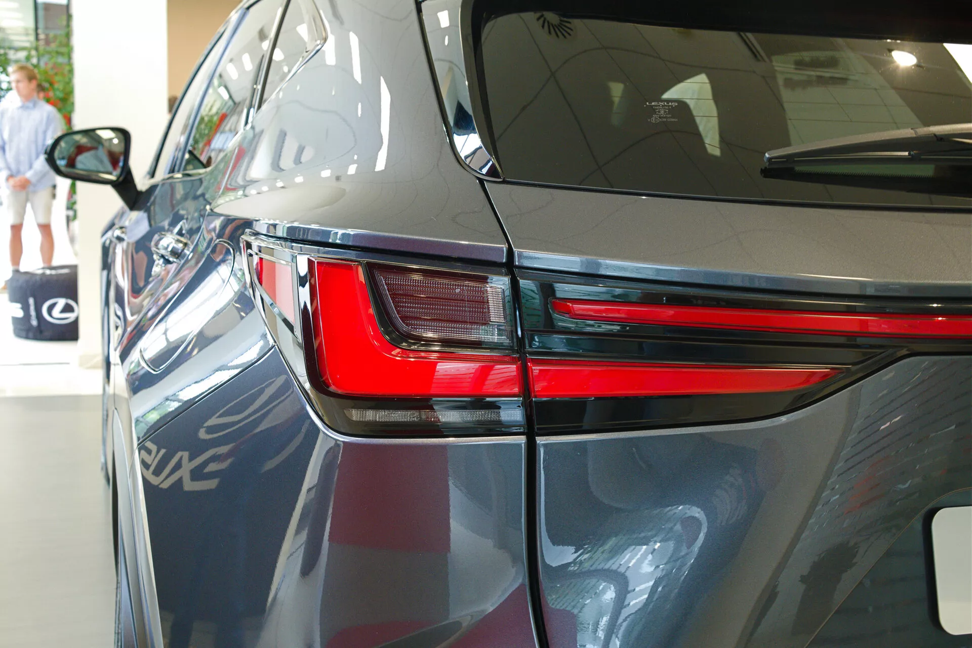 Närbild på 2021 Lexus NX från en bakre trekvartsvinkel som framhäver den eleganta LED-baklyktan, en del av bakre kvartspanelen och en del av bagageluckan. Bilden visar bilens blanka mörka lack, aerodynamiska karosslinjer och antyder en inomhusutställningsmiljö med reflektioner av ett annat fordon, arkitektoniska element och en person som går i bakgrunden.