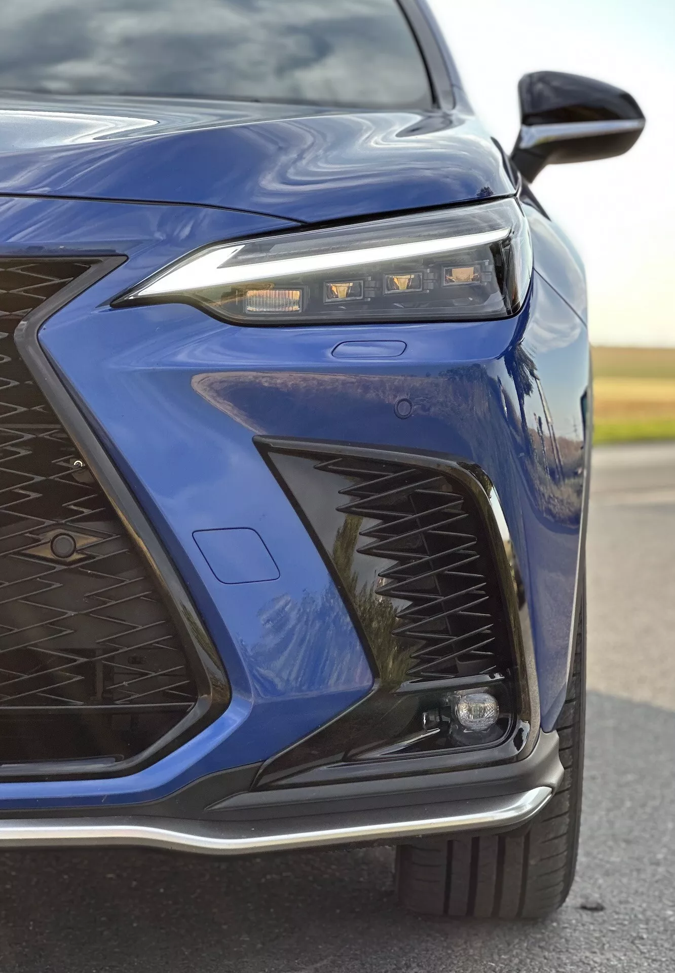 Närbild av främre vänstra hörnet på en Lexus NX från 2021 i mörkblått som framhäver avancerad LED-strålkastardesign, invecklat grillmönster, sportig frontstötfångare och eventuella adaptiva belysnings- och aerodynamiska funktioner med en glänsande metallisk finish