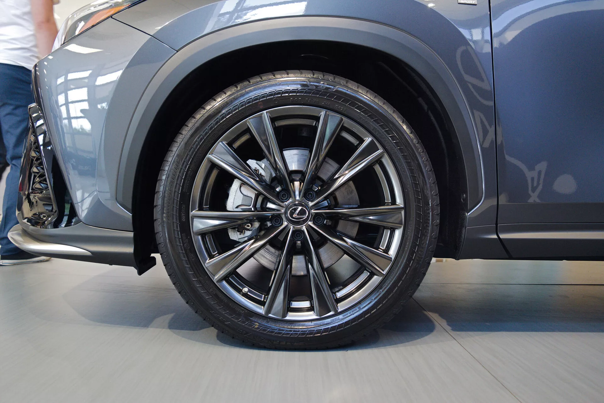 Närbild i lågvinkel på förarsidans framhjul av en Lexus NX 2021, som visar en legeringsfälg med fler-ekrade design och tillverkarens logotyp. Lågprofils-däcket framhäver sportig prestanda. Stänkskärm över hjulet är metallisk och något utbuktande, vilket antyder sportighet. Sidopanelen visar en elegant, aerodynamisk kontur. Bild tagen inomhus, med närfokuserad perspektiv på hjulet och den främre sidopanelen.