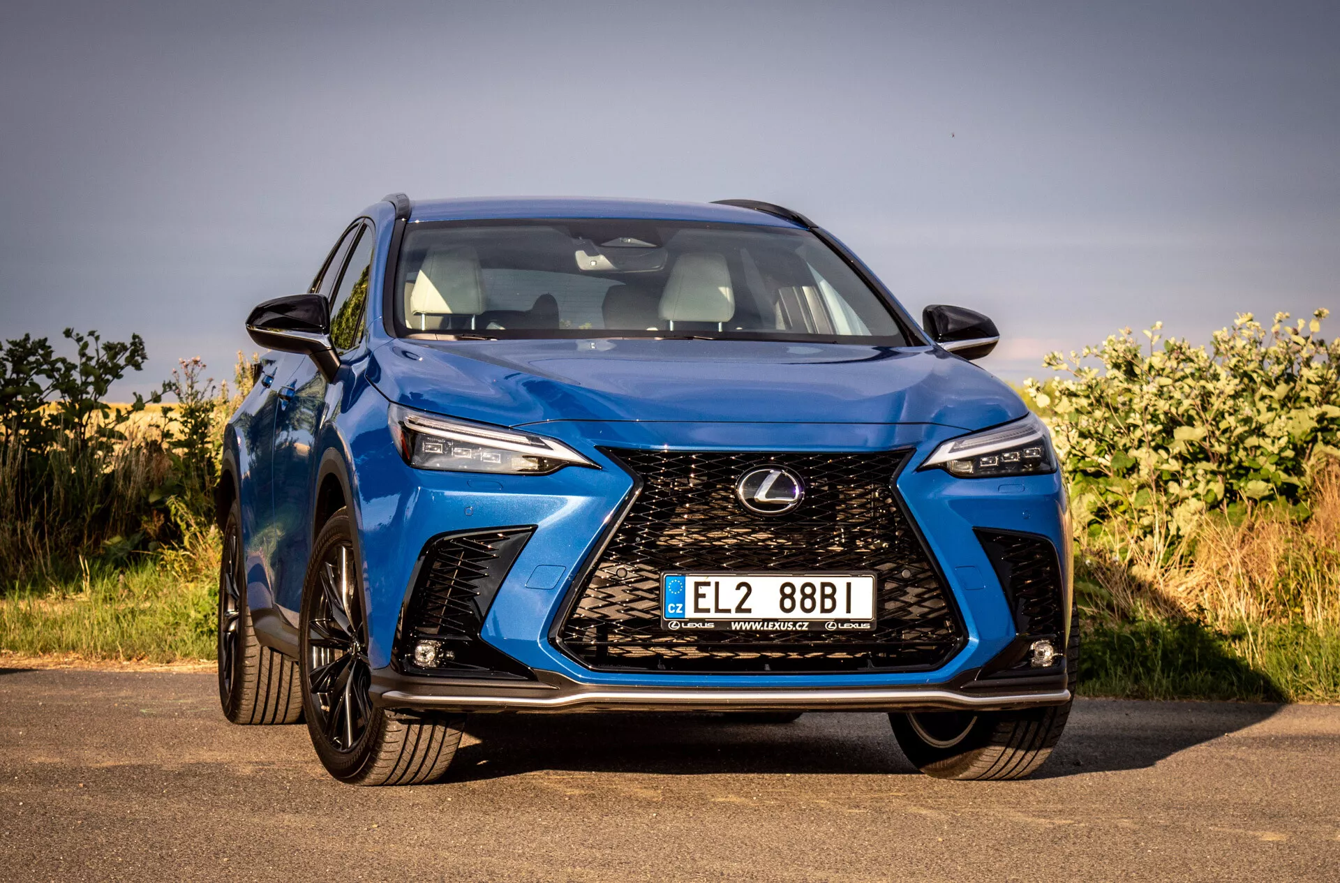 Frontvinkel vy av en blå Lexus NX 2021 SUV med fokus på dess intrikata spindelgrill, vinklade LED-strålkastare, glansig finish och aerodynamisk design, parkerad utomhus under klar himmel