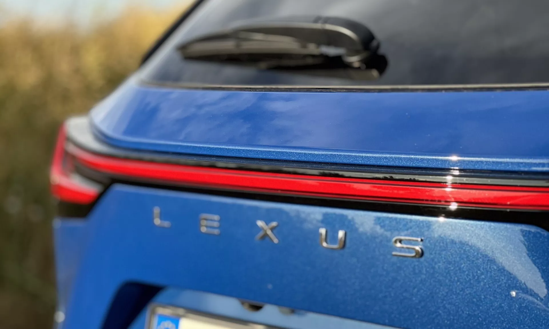 Närbild av den bakre varumärkesområdet för Lexus NX 2021 med en silverfärgad Lexus-badge, ett rött ljusstråle ovanför namnet, på en metallisk blå kropp under dagsljusförhållanden