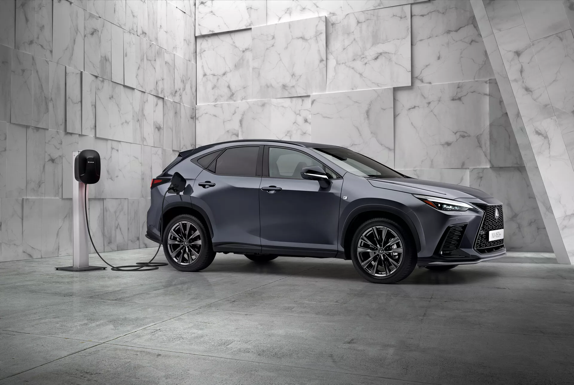 Denna bild visar 2021 Lexus NX, en modern kompakt SUV med en elegant och sportig design, fotograferad från tre fjärdedels framåtriktad vinkel. Fordonets vänstra sida och delvis dess front är synliga och positionerade diagonalt i en minimalistisk miljö prydd med stora ljusa marmorkakel, vilket antyder en premiumatmosfär. Lexus NX har karakteristiska LED-strålkastare och en framträdande frontgrill, vilket betonar dess sofistikerade natur. Synliga aerodynamiskt formade sidobackspeglar, komplexa fälgar med flera ekrar och infällda dörrhandtag framhäver den sportiga och avancerade designen. En viktig detalj är att bilen är ansluten till en elbilsladdningsstation, vilket indikerar att det är ett elfordon eller plug-in hybrid. Framskärmen har en laddport, vilket visar dess elektriska kapacitet. Denna statiska bild fokuserar på att visa bilens design och teknologiska funktioner i en lugn, balanserad komposition med mycket negativt utrymme, vilket symboliserar exklusivitet och innovation inom bildesign.