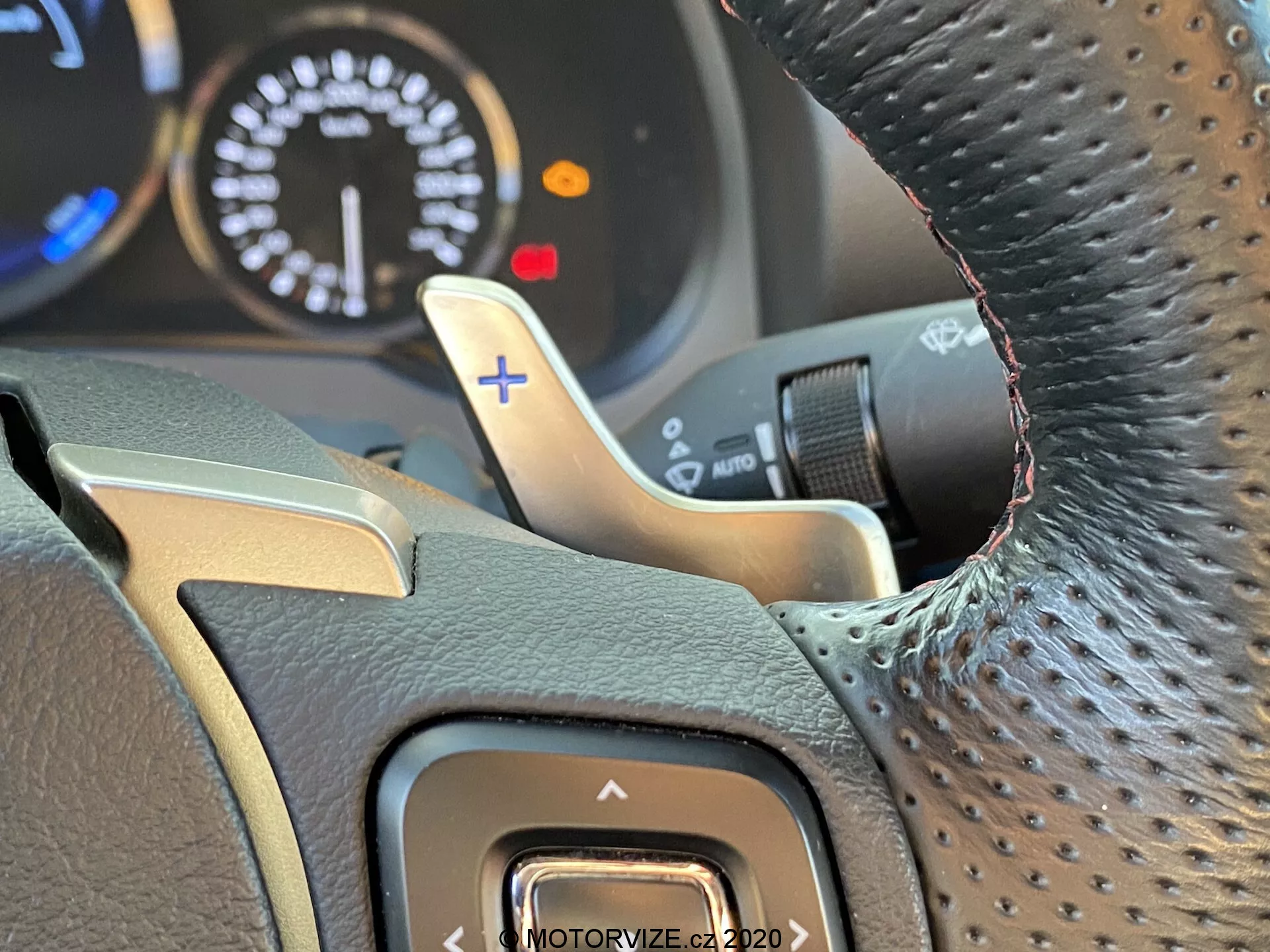 Närbild från förarens perspektiv av Lexus RC 2016 ratt med texturerat grepp, möjligen av läder, kromaccent, knapp för förarassistans på vänster sida, metallisk växelpaddel bakom högra sidan av ratten med '+'-tecken för manuell växling uppåt, och delvis synlig instrumentpanel som antyder sportmotor med varvräknare som når 8000 RPM; inre fokus med suddig bakgrund som framhäver detaljerna i ratten och växelpaddeln.