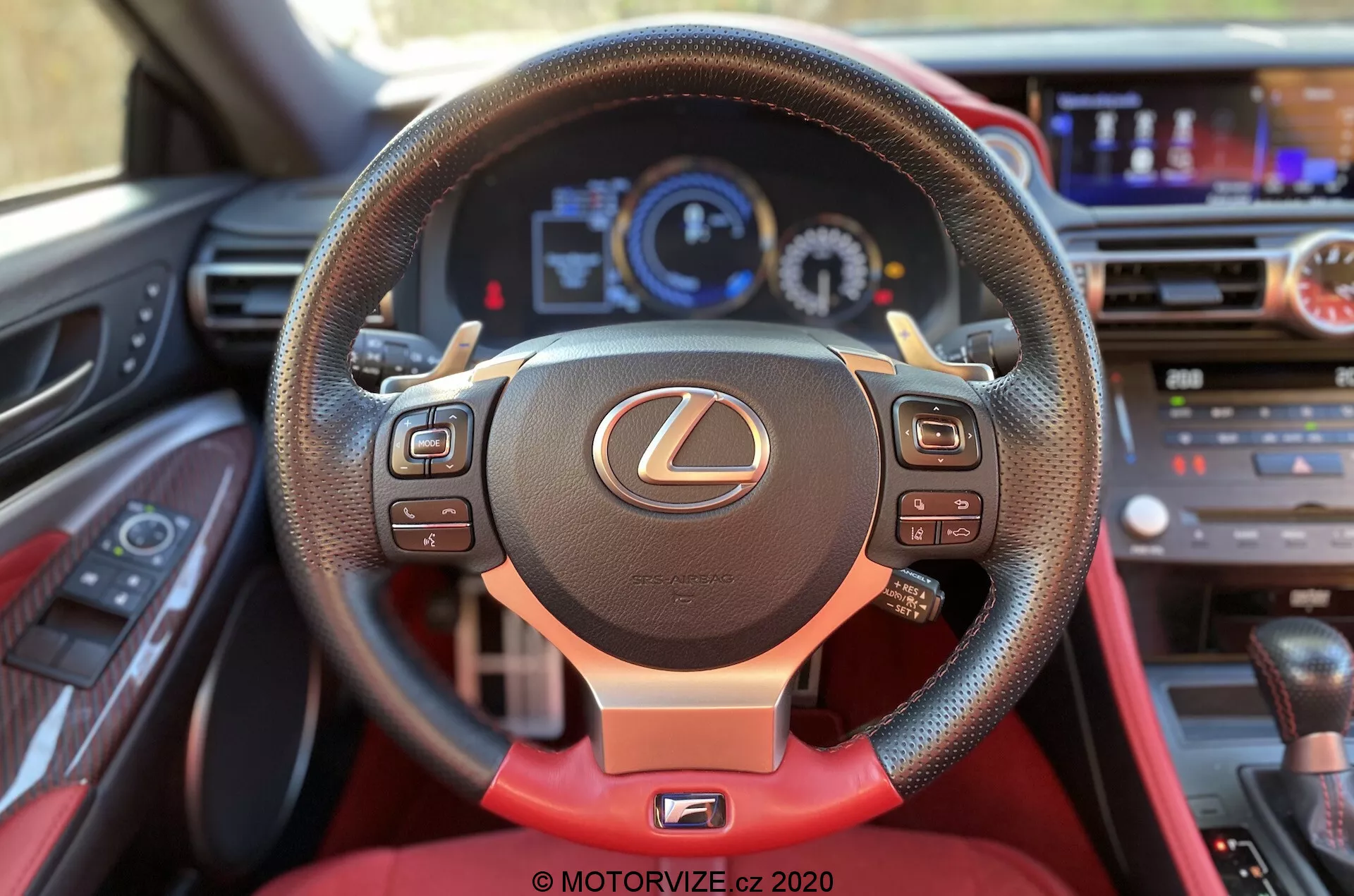 Interiörvy från förarsätet i en Lexus RC 2016, med fokus på ratten med Lexus-emblemet. Den tre-ekrade ratten har multifunktionskontroller, läder med röda sömmar och en röd accent på den nedre eken. Det digitala instrumentklustret bakom ratten visar en hastighetsmätare, varvräknare och en multifunktionsdisplay. Den övre delen av instrumentbrädan innehåller ett infotainmentsystem och klimatkontrollknappar. Nedanför instrumentbrädan syns en del av mittkonsolen med växelväljaren.