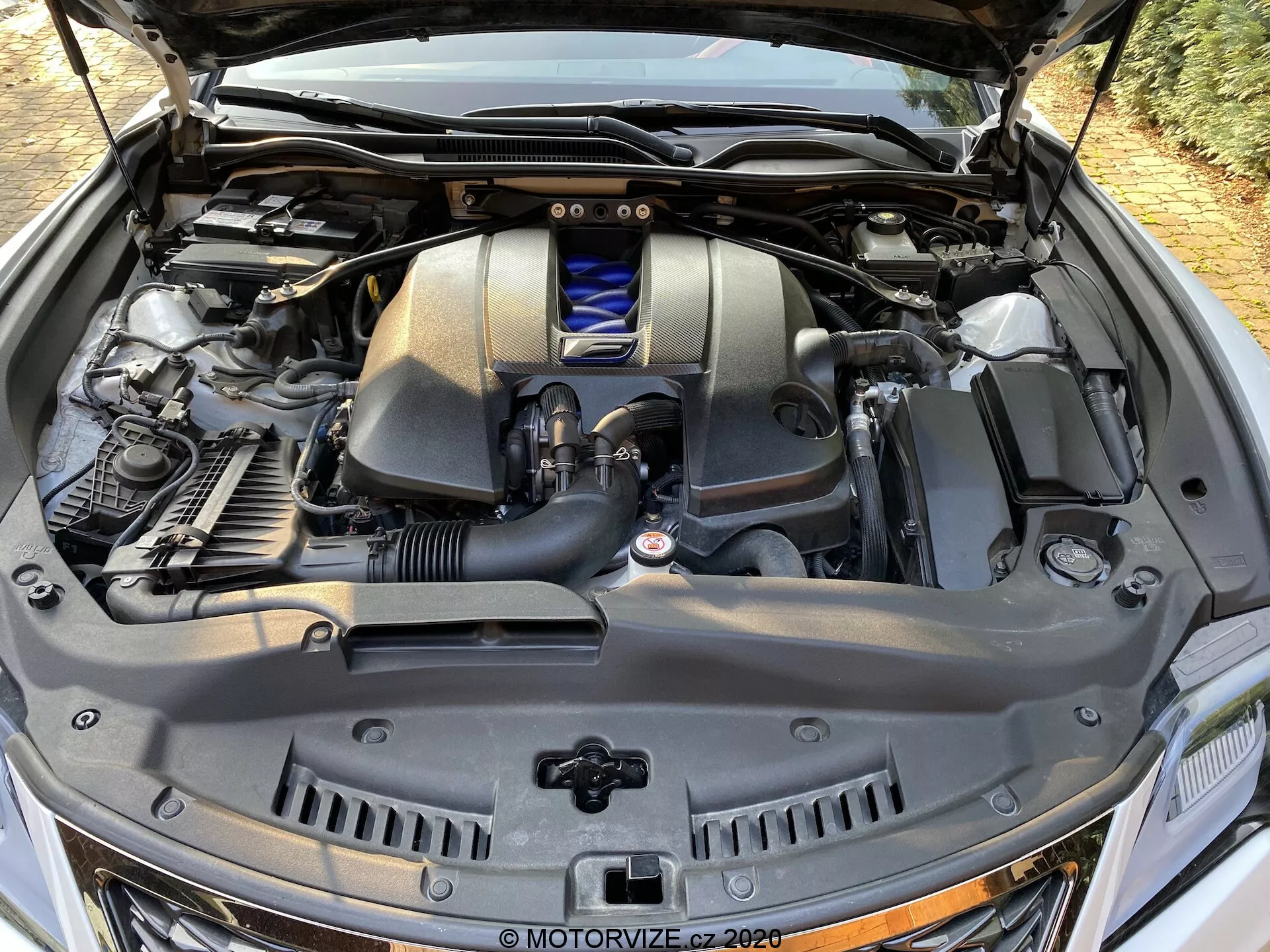 Detaljerad närbild av motorutrymmet på en 2016 Lexus RC från en framifrån-höger vinkel, som framhäver motorlocket med emblem, luftintagssystemet, vätskebehållarna med blått lock, metalliska och gummierade slangar, övre delen av kylaren samt strukturella komponenter mot bakgrunden av brandväggen och bilens elektroniska moduler.