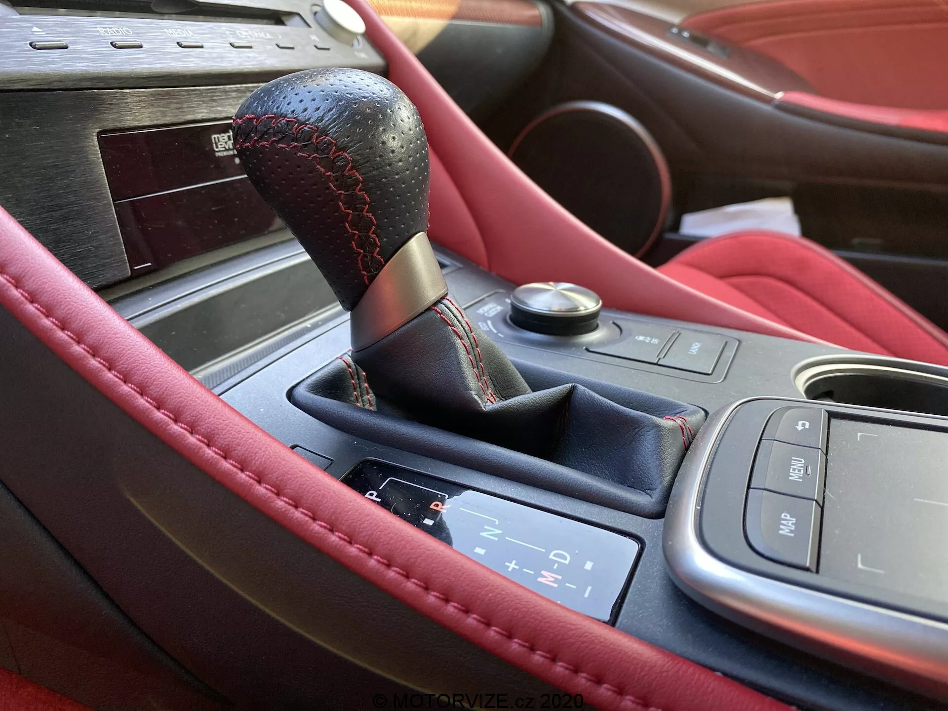 Närbild av mittkonsolen i interiören på en 2016 Lexus RC, som visar en växelspak i svart läder med röda sömmar, elektronisk växelpanel för automatisk växellåda, multimediekontrollgränssnitt, motor start/stopp-knapp och lyxig träpanel. Funktionerna inkluderar premium lädermaterial och troligtvis mugghållare, vilket antyder en sportig och lyxig design.