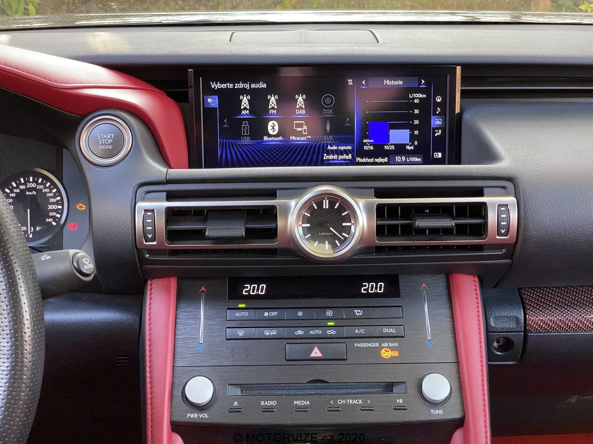 Bilden visar interiören i en Lexus RC 2016 från perspektivet av en av framsätena och visar en modern, multifunktionell pekskärmsdisplay i mitten av instrumentbrädan. Ovanför skärmen sitter en elegant analog klocka. Instrumentbrädan har vertikala luftventiler på varje sida av skärmen, justerbara horisontella spjäll för luftflöde, och många fysiska kontroller och knappar för klimatkontroll, stolsvärme, media- och radiofunktioner. Den lilla klimatdisplayen indikerar ett tvåzons klimatsystem. Blandningen av röda läderdetaljer och svart texturerat material ger en sportig, högkvalitativ estetik, medan omgivningsbelysningen ger en förfinad touch.