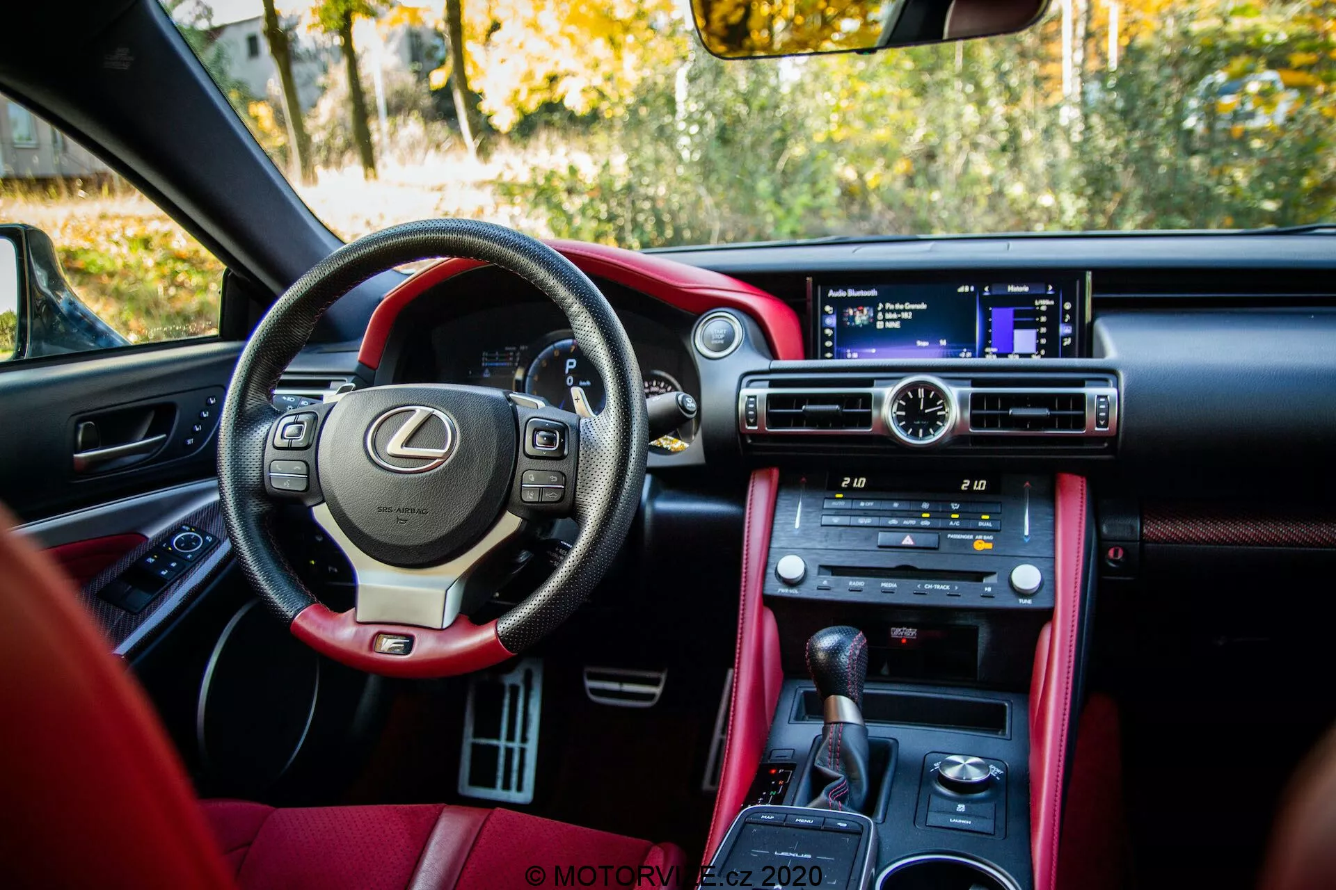 Interiörvy av en parkerad Lexus RC 2016 från bakre passagerarsätets perspektiv. Bilden visar en lyxig frontkabin med en multifunktionell läderklädd ratt med paddlar, antydande om en sportig karaktär. Den breda instrumentbrädan har en icke pekskärmsinfotainmentskärm med en fjärrkontroll pad på den läderfodrade växelspaken på mittkonsolen. Fysiska knappar för klimatanläggningen och sätesuppvärmning/kylning är synliga, tillsammans med körlägesval som Eco, Normal och Sport. Den tvåfärgade interiören, med en kombination av mörka färger och kontrasterande rött läder, badar i naturligt dagsljus och visar en stillastående utomhusmiljö med löv som antyder en höstscen.