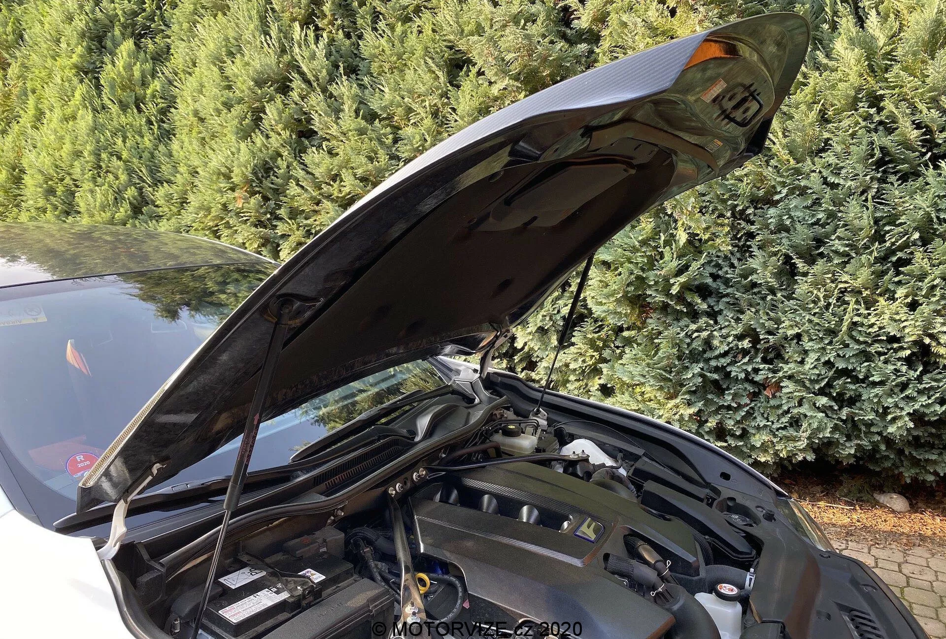 Detaljerad närbild av framsidan av en Lexus RC 2016 med öppen motorhuv, som visar en delvis skymd motorrum från en sned vinkel. Motorhuven är öppen nästan 90 grader, vilket visar den ergonomiska gångjärnsmekanismen lämplig för underhåll. Olika motorkomponenter, vätskebehållare och strukturella delar är synliga, även om specifika motorteknologier inte är tydligt synliga. Bildens bakgrund inkluderar barrträd, vilket antyder en utomhusplats.