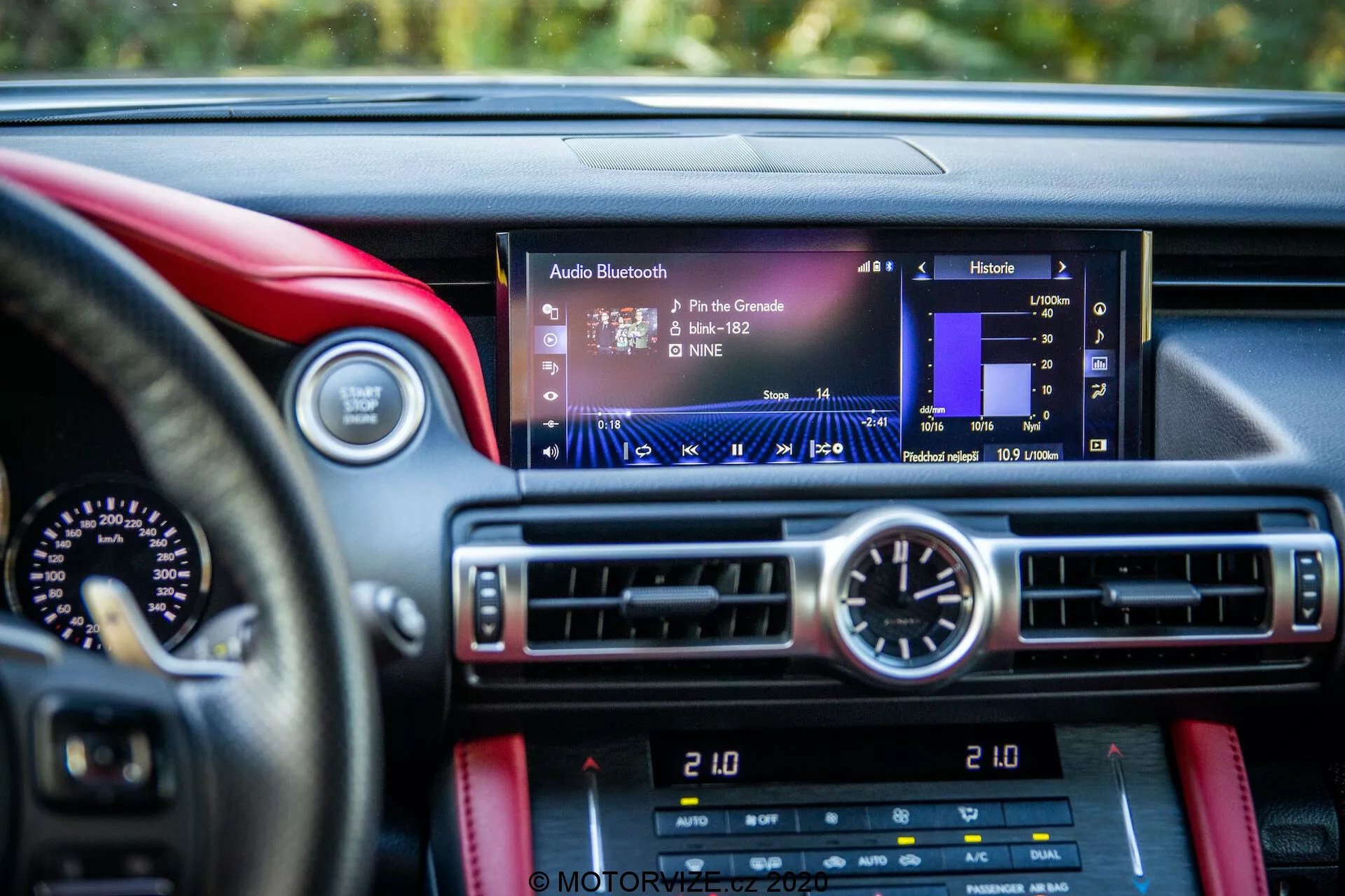 Närbild från förarsätet i en Lexus RC 2016 som visar den moderna infotainmentskärmen med element som spårnamn, artist och ljudnivåer, centrerad i ramen. Under skärmen finns klimatkontroller och en rund analog klocka. Instrumentpanelen har en lyxig eller sportig design med kontrasterande material, möjligen läder med röda sömmar mot en mörkare bakgrund. Delvis synliga är hastighetsmätaren och varvmätaren, tillsammans med ytterligare bilkontroller under klimatkontrollerna.