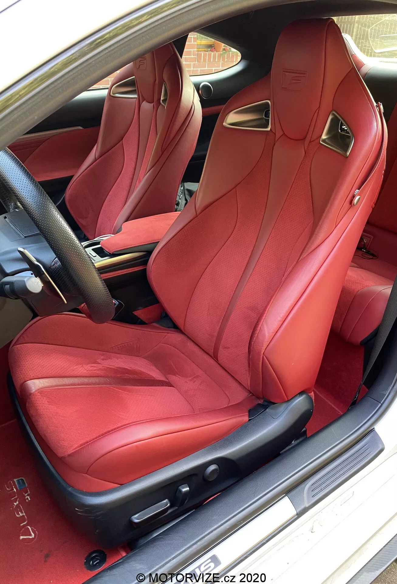 Interiörvy av en 2016 Lexus RC, som visar den lyxiga röda lädersätet för passagerare fram med sportiga sidostöd och integrerat nackstöd, sett från förarsidan. Sätet har intrikat sömnad och elektriskt justerbara kontroller, kompletterade med metalliska accenter. Bildörren är öppen, vilket avslöjar det märkesbelagda tröskelplattan, med en glimt av den bostadstegelbeläggningen utanför.