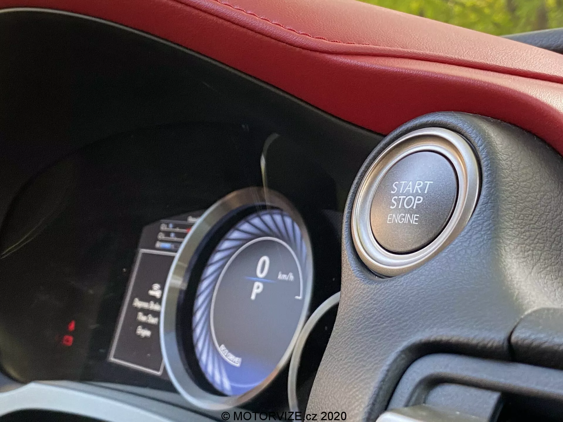 Närbild av Lexus RC 2016 interiör från förarens perspektiv, som visar en digital-analog instrumentpanel med en hastighetsmätare som visar '0 km/h' och en bränslemätare. Fokus ligger på det moderna tändningssystemet med tryckknapp märkt 'START STOP ENGINE' omgivet av en metallring, vilket indikerar ett premiumfordon med nyckellös motorstart. Den välbelysta hytten har inslag av röd klädsel och avancerad fordonsteknik.