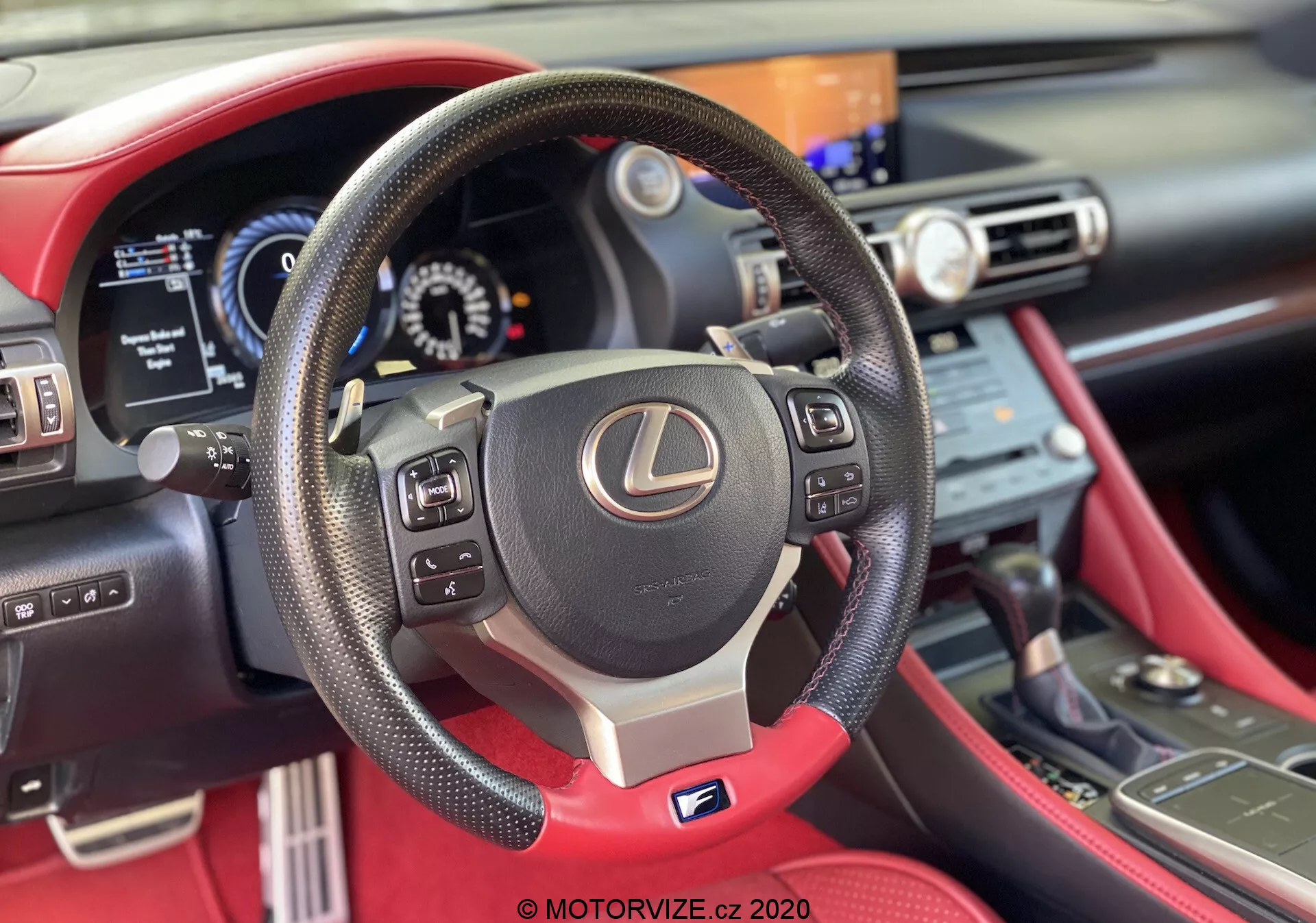 Förstapersonsvy av interiören i 2016 års Lexus RC, med fokus på en centralt placerad treekrad läderklädd ratt med kontrollknappar, växelpaddlar och en silver 'F' Sport-emblem. Delvis synlig digital instrumentpanel och mittkonsol med infotainmentskärm och klimatkontroller. Slående röd läderinredning tillför en premiumkänsla.