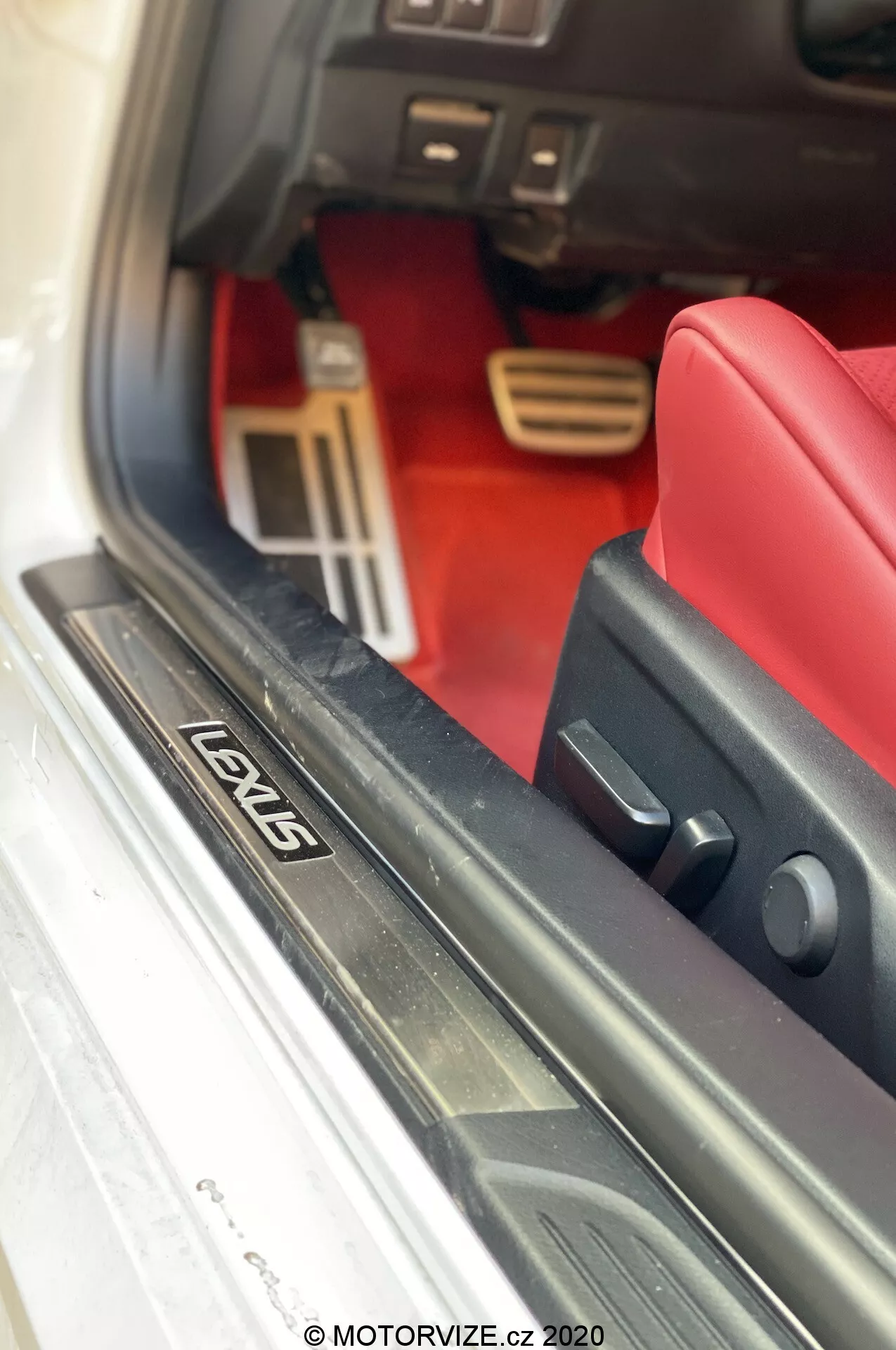Närbild av insidan av en Lexus RC 2016, med metalliska dörrtröskeln med 'LOVIS' plåt, rött mattat golvområde, sportiga aluminiumspeddaler, nedre instrumentpanelen och svart gummitätning på dörren. Foto taget av MOTORVIZE.cz 2020.