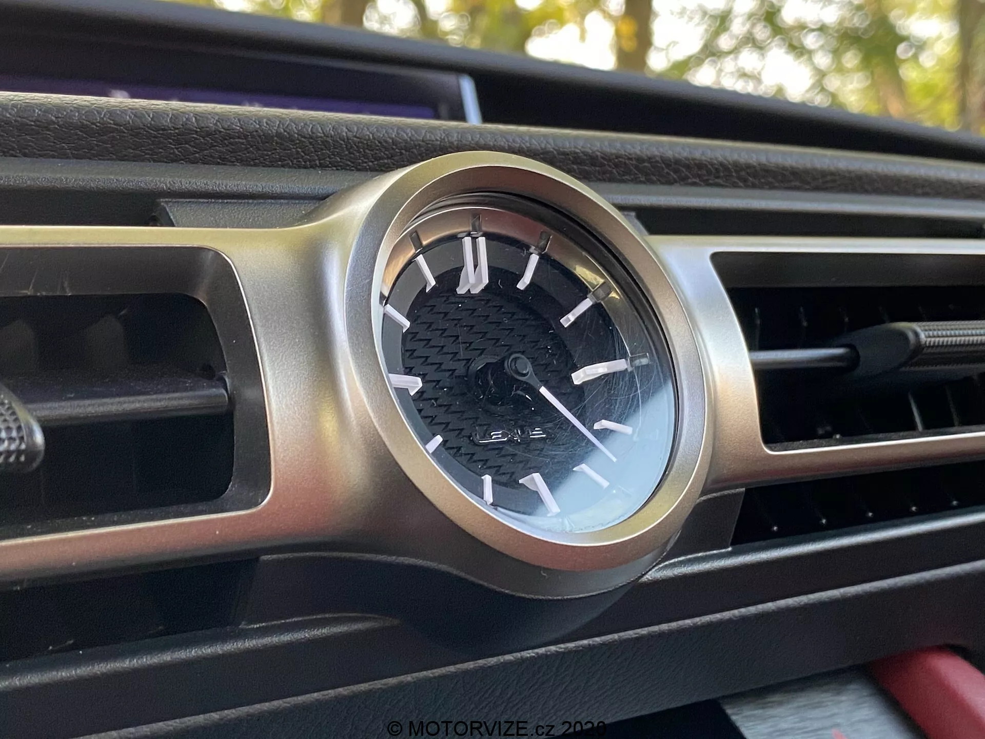Närbild, något vinklad vy av en cirkulär analog klocka på Lexus RC:s instrumentpanel från 2016, innesluten i ett borstat metallhölje. Klockan har en svart bakgrund med vita markeringar, timsiffror och minutmarkeringar, placerad i mitten av en horisontellt utsträckt luftventil. Instrumentpanelen har ett texturerat svart mönster, möjligen av kolfiber. Bakgrundselementen är suddiga, vilket fokuserar uppmärksamheten på klockans detaljerade design.