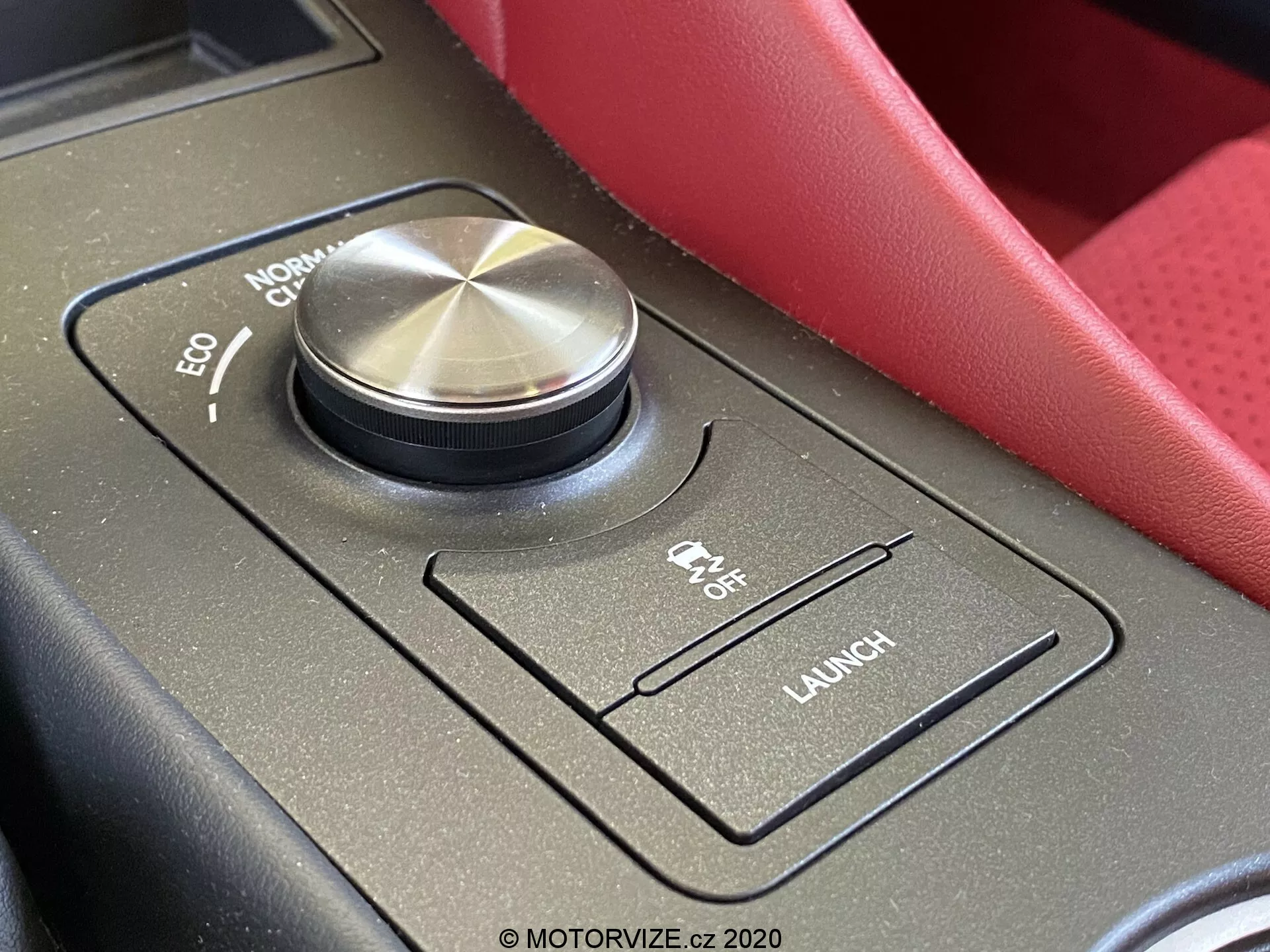 Närbild av området för körlägesväljaren inuti en Lexus RC 2016, som visar en stor flerlägesväljare med bearbetad metallfinish och räfflade kanter. Väljaren visar lägena 'ECO,' 'NORMAL,' 'COMFORT' och ett delvis skymt sportläge. Runt väljaren finns ytterligare kontroller på en matt svart yta, inklusive en 'CTX OFF' växel för antisladdsystemet och en 'LAUNCH'-knapp för startkontroll. Bilden fångar mittkonsolen från förarsätets perspektiv och framhäver de premium- och prestandaorienterade interiörfunktionerna.