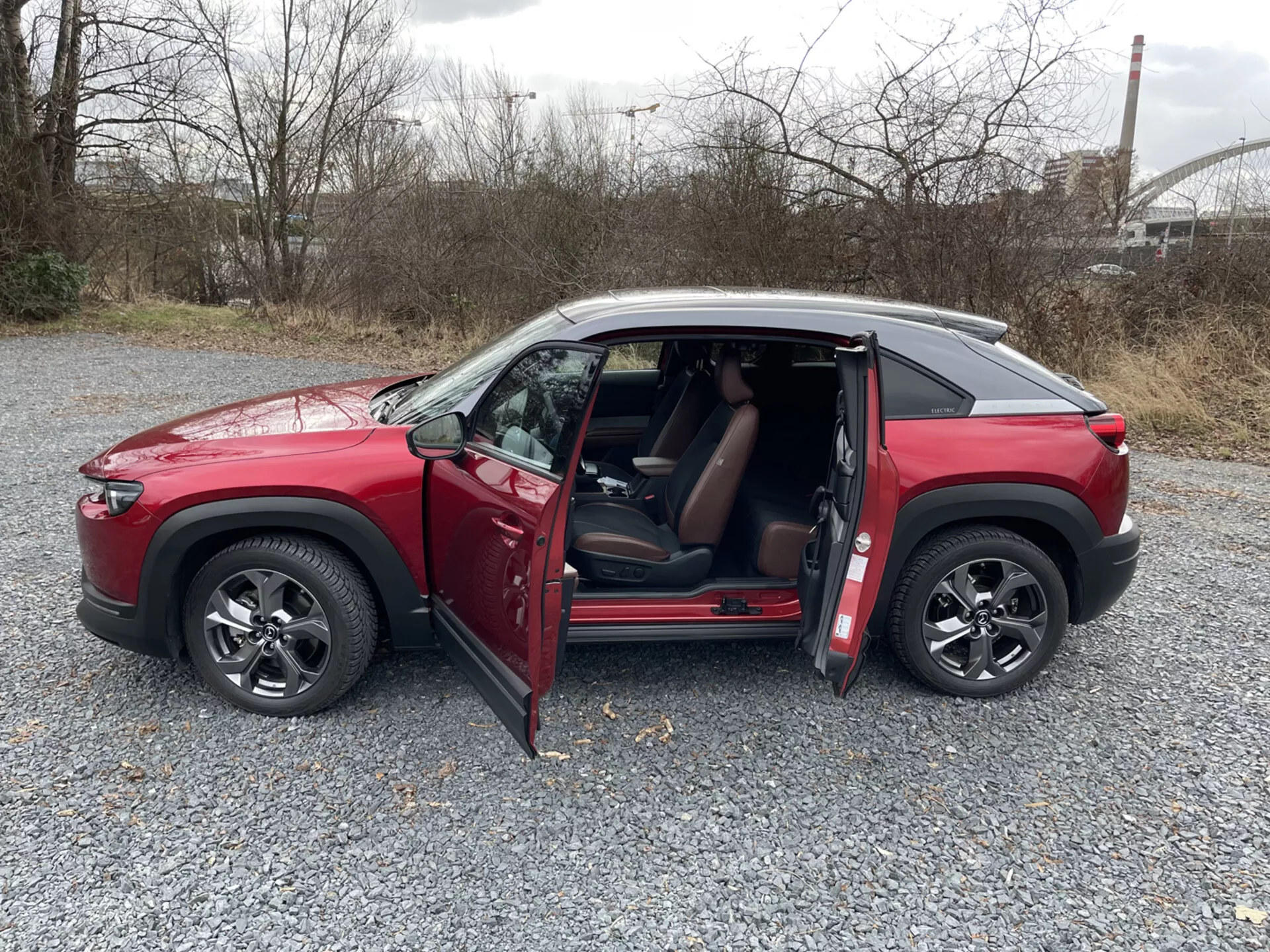 2020 Mazda MX-30 röd fyrdörrars SUV i sidoprofil på grus med öppna dörrar, flerstråliga lättmetallfälgar, tonade fönster och kontrastfärgat tak mot en bakgrund av industri och grönska.