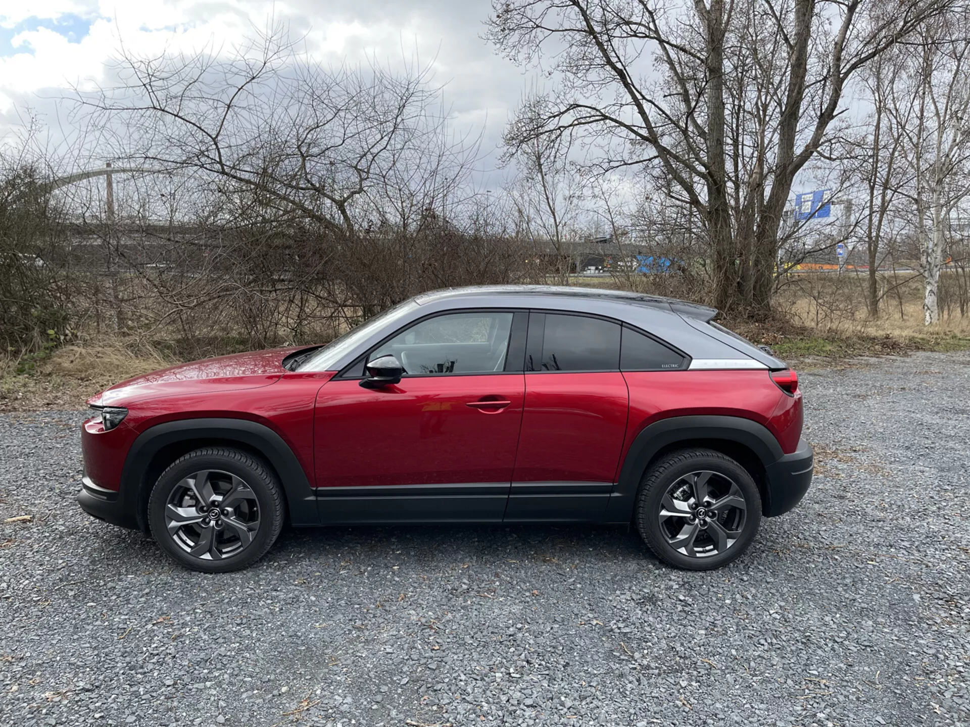Sidoprofil av en röd Mazda MX-30 crossover SUV 2020 på grus med glest vegetation och molnig himmel, visar hög midjelinje, kupéliknande sluttande taklinje, dynamisk karaktärslinje, fälgar med flerspakiga legeringar med svart plastklädsel och möjliga avancerade tekniska funktioner som ADAS, LED-dagsljus, nyckelfritt inträde, däcktrycksövervakningssystem TPMS och ett infotainmentsystem.