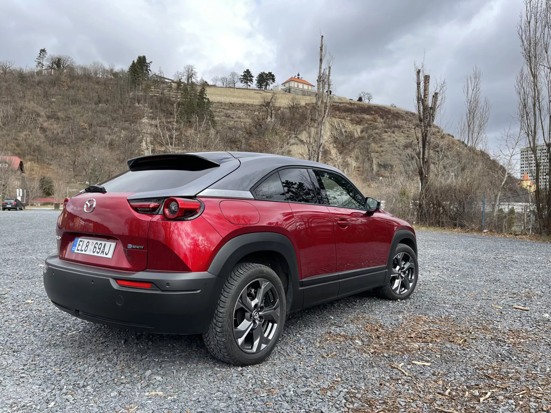 2020 års Mazda MX-30 modern crossover parkerad på grus, som visar dess sportiga tvåfärgade röd och svarta design, LED-bakljus, bakspoiler och flerstråliga lättmetallfälgar mot en bakgrund av natur och arkitektur